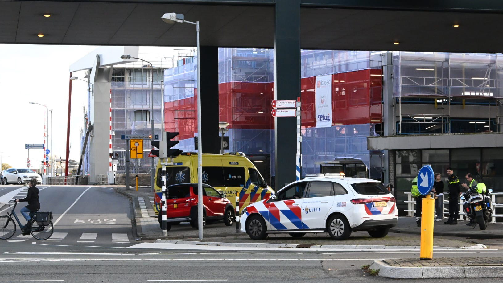 Een straatbeeld met een ambulance en een politieauto naast een brug, een fietser voorbijrijdend op een fietspad, en enkele mensen, waaronder agenten, staan op de achtergrond bij een bouwlocatie.