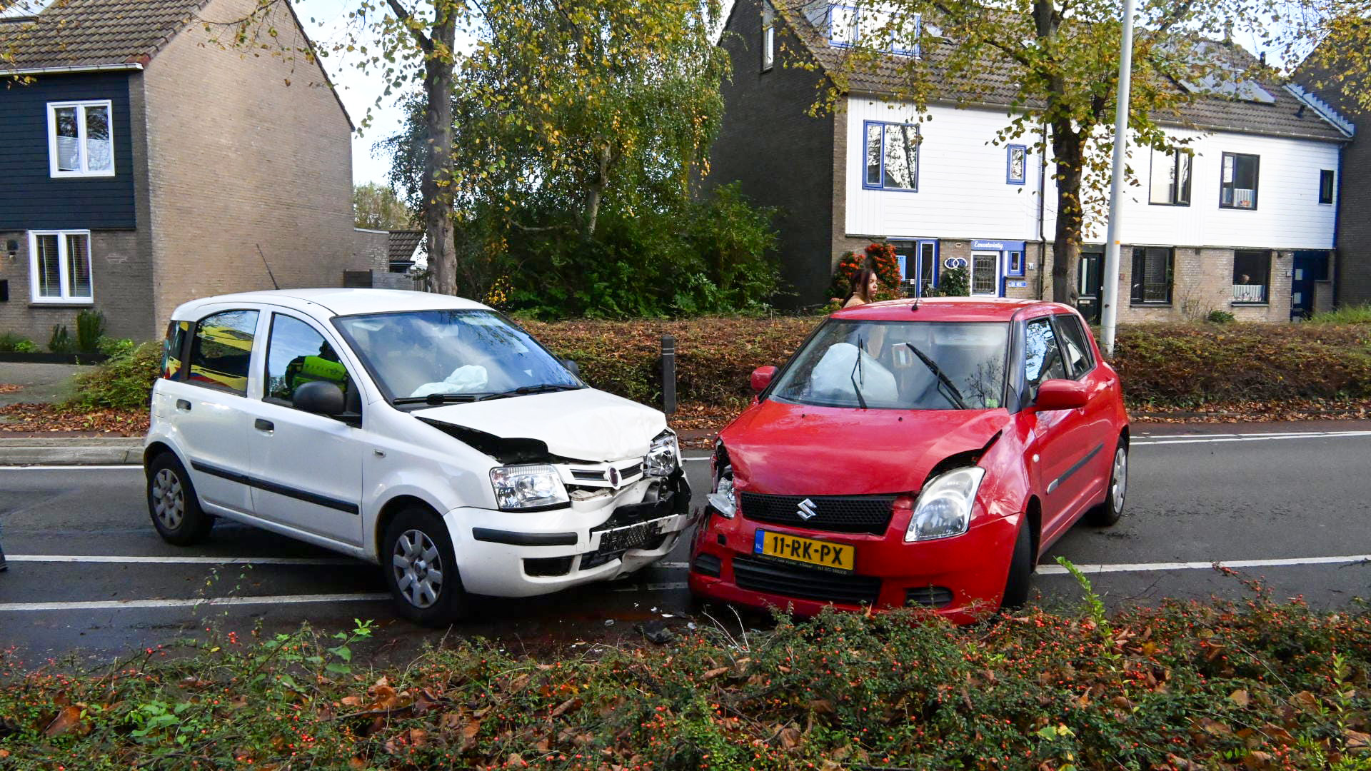 Een botsing tussen een witte auto en een rode auto op een rustige straat, omgeven door woonhuizen.