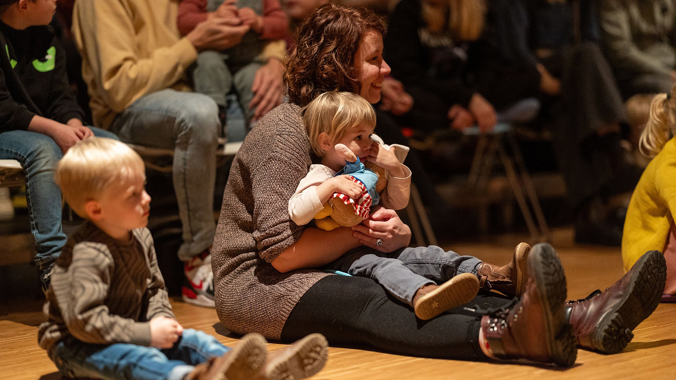 Een vrouw zit op de grond met een kind op schoot dat een teddybeer vasthoudt, omringd door andere kinderen zittend op een houten vloer.