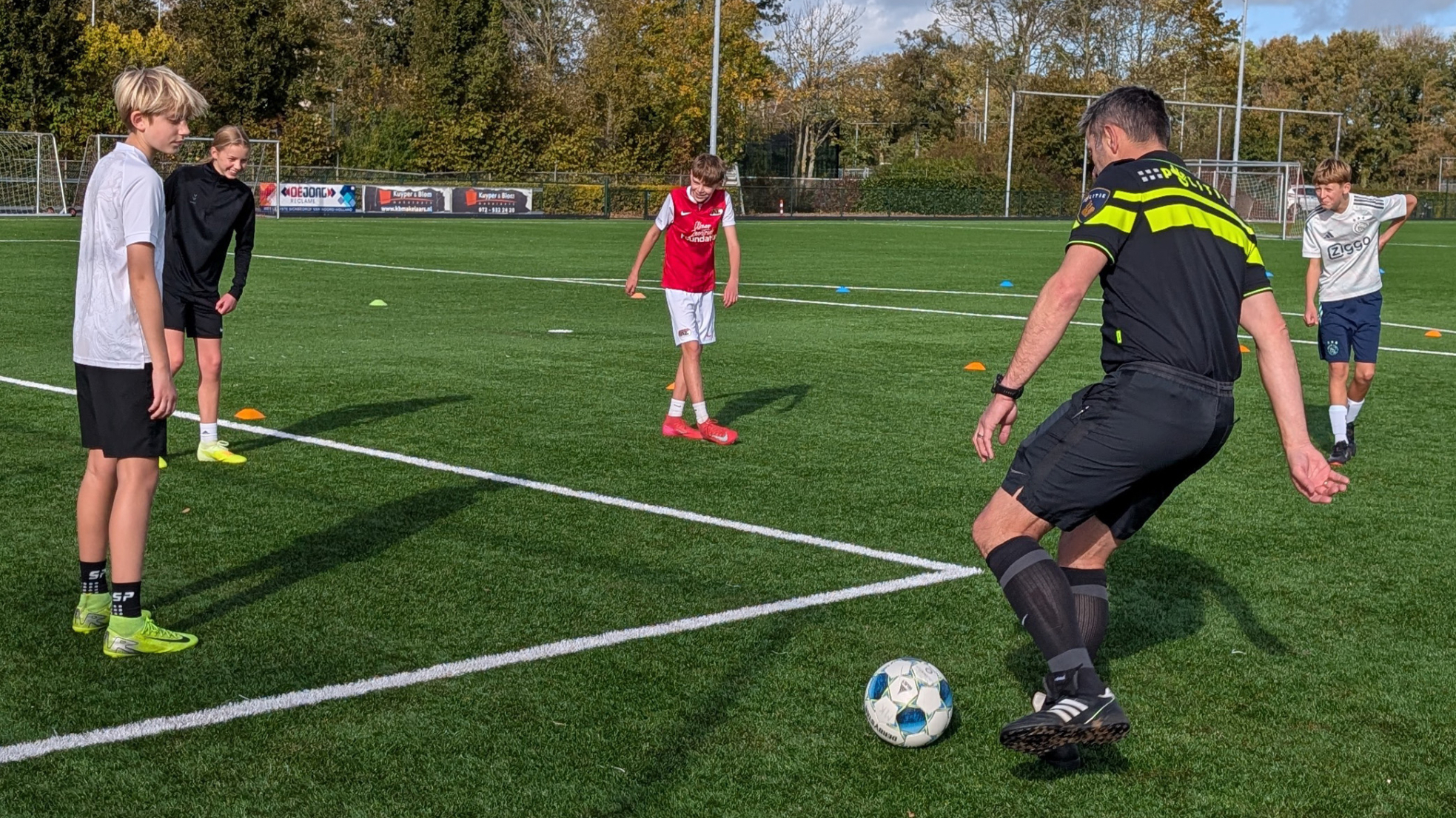 Een groepje voetballers op een veld, terwijl een volwassen man in sportkleding de bal dribbelt, omringd door vier jongeren die aandachtig kijken. Op de achtergrond zijn doeltjes en bomen zichtbaar.