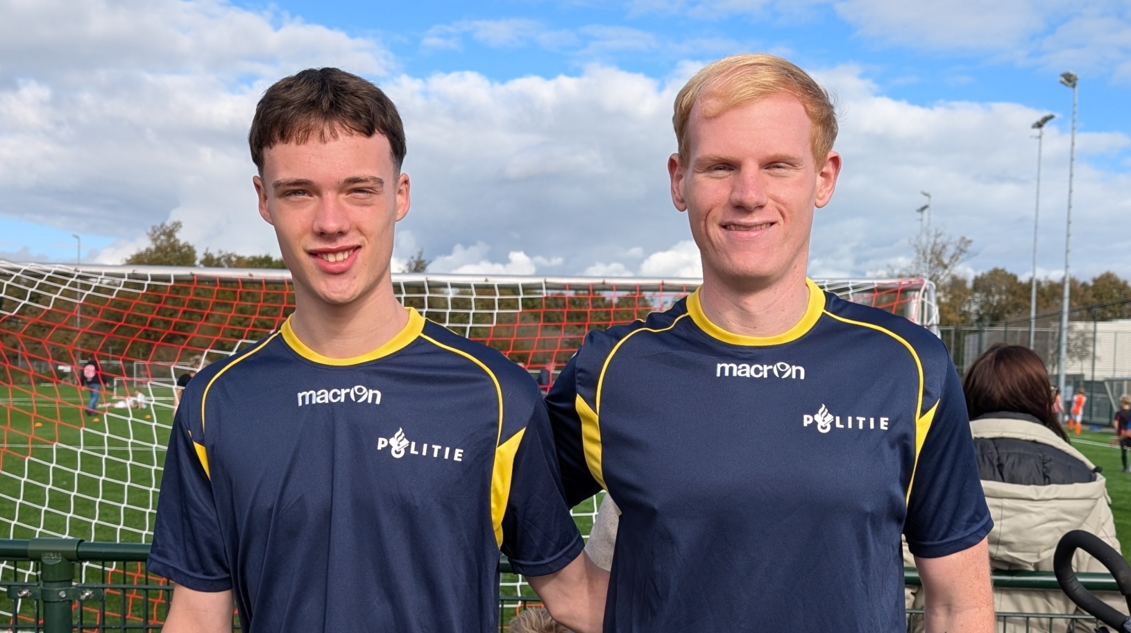 Twee mannen in donkerblauwe sportkleding met het opschrift 'Politie' poseren voor een voetbaldoel op een veld, omringd door mensen en een fiets vooraan in beeld.