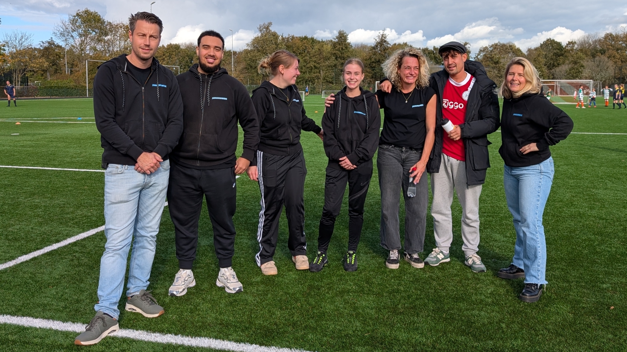 Groep mensen, deels in zwart sportkleding gehuld, poseren samen op een voetbalveld; op de achtergrond zijn juveniele spelers actief bezig met een wedstrijd.