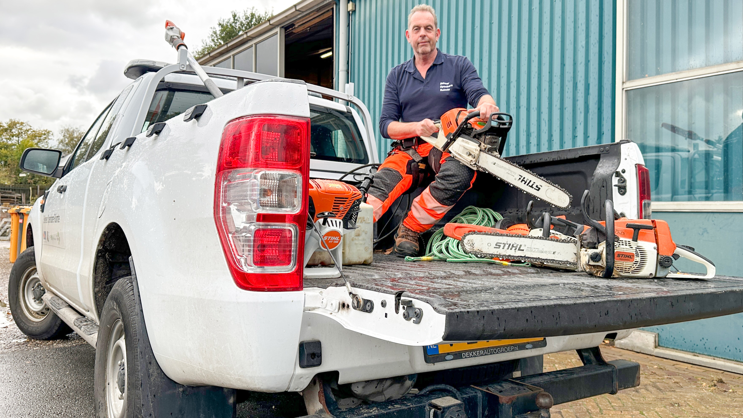 Man in work attire seated on the tailgate of a white pickup truck, holding a chainsaw and surrounded by tools; the truck is parked outside a building with a blue wall.
