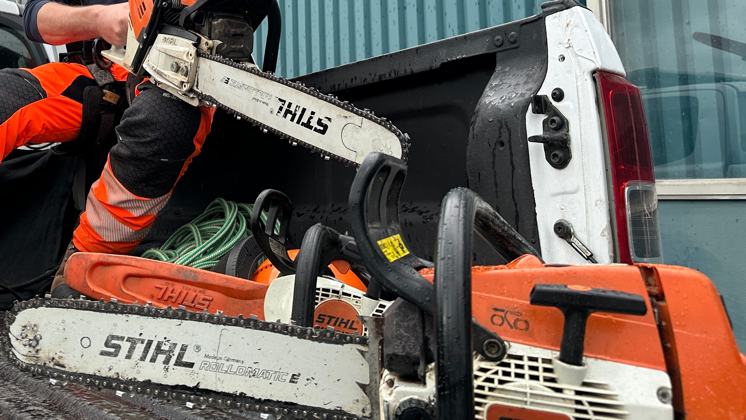 Een persoon in oranje veiligheidskleding houdt een Stihl kettingzaag bij een open laadbak met meerdere kettingzagen en een groene haspel.