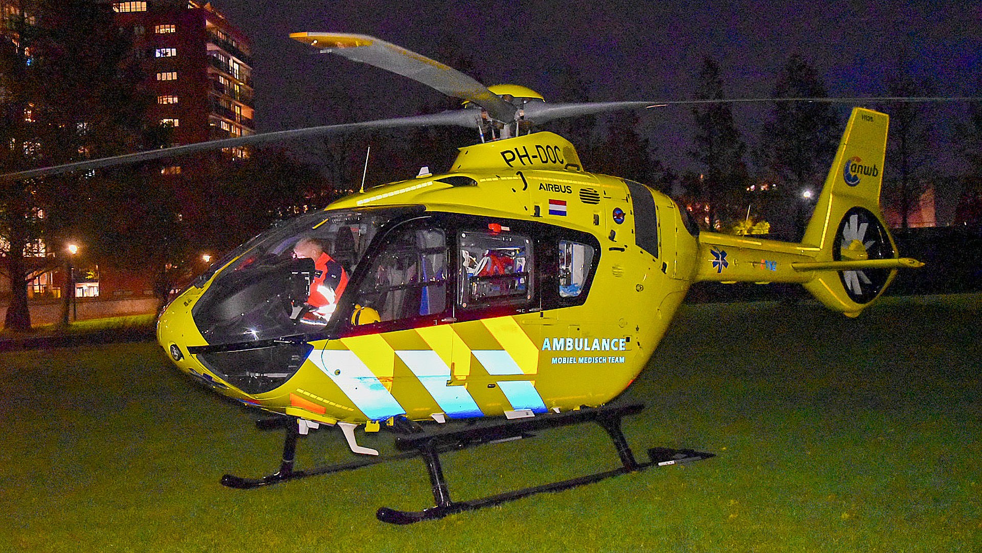 Een gele traumahelikopter van de ANWB, met het opschrift 'AMBULANCE Mobiel Medisch Team', geparkeerd op een grasveld bij nacht met een stadsachtergrond.