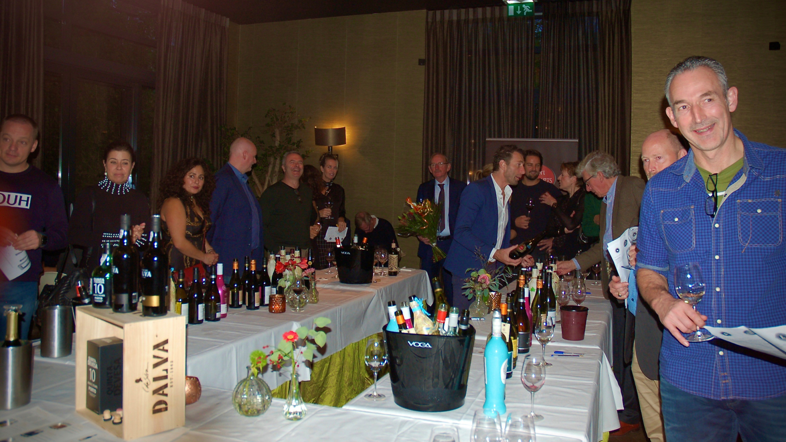 Mensen staan rond een tafel vol met wijnflessen tijdens een proeverij in een zaal.