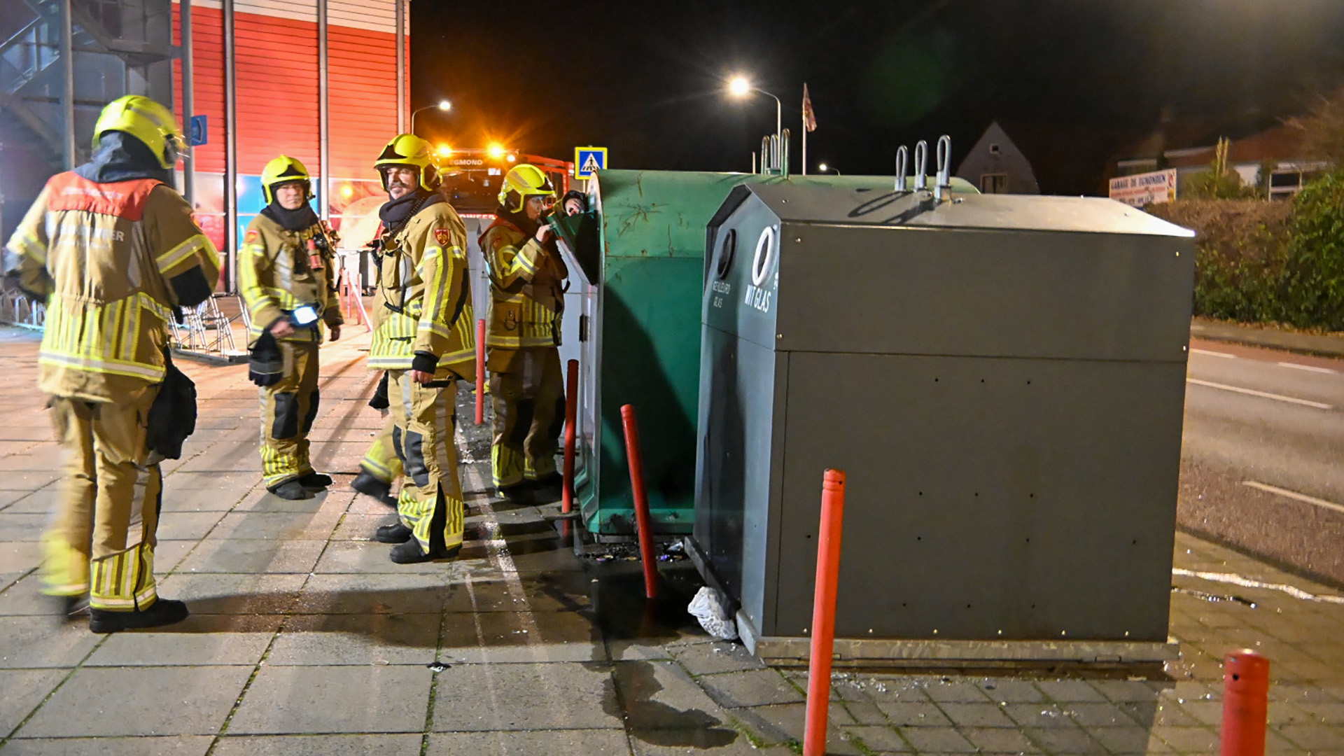 Brandweermannen in gele uniformen staan 's nachts naast een glascontainer op een stoep. Er is een brandweerauto met felle lichten zichtbaar op de achtergrond.