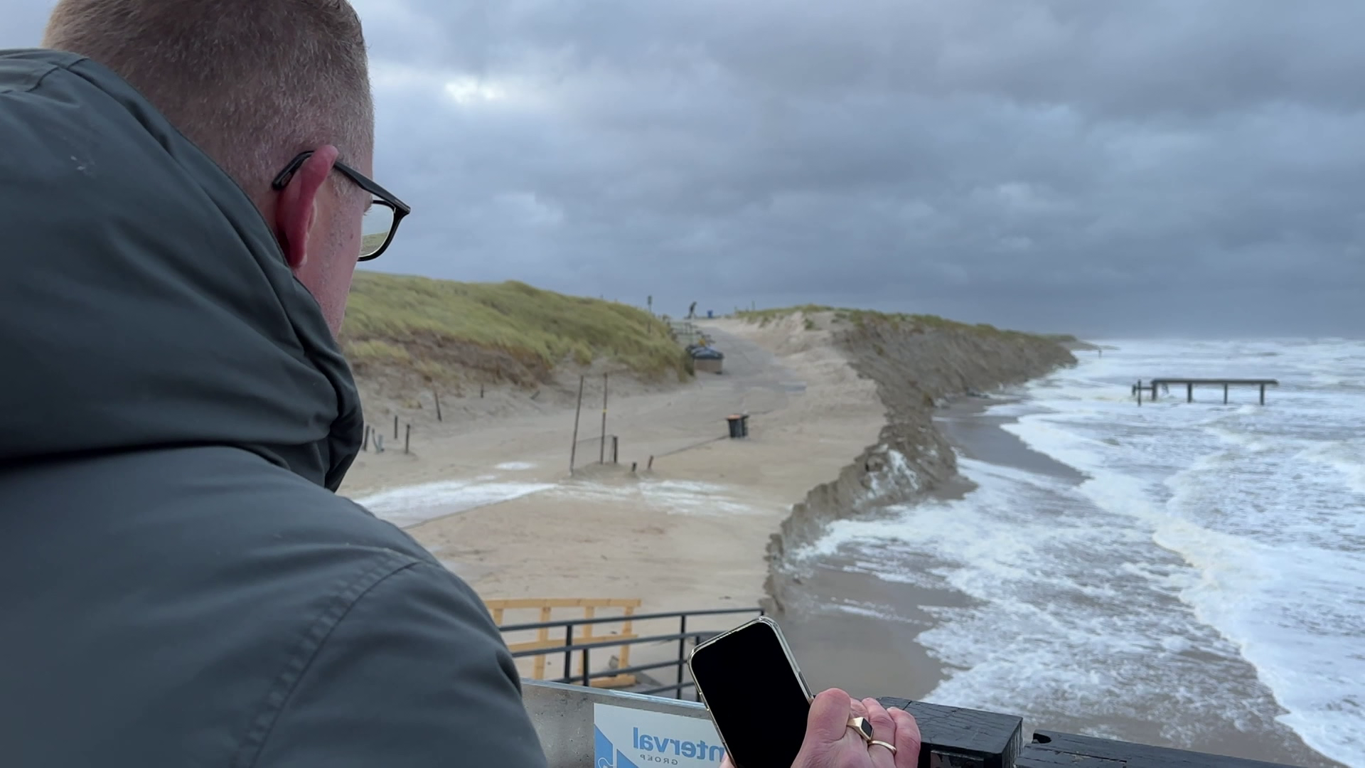 Man met bril en jas kijkt naar een zandstrand en ruwe zee, met een houten pier aan de rechterkant op een bewolkte dag.