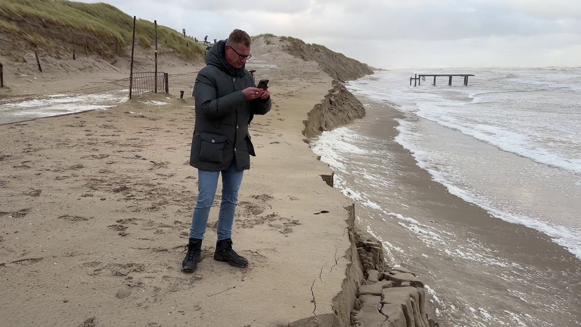 Man in winter clothing stands on eroded beach near the sea, taking a photo with a smartphone.
