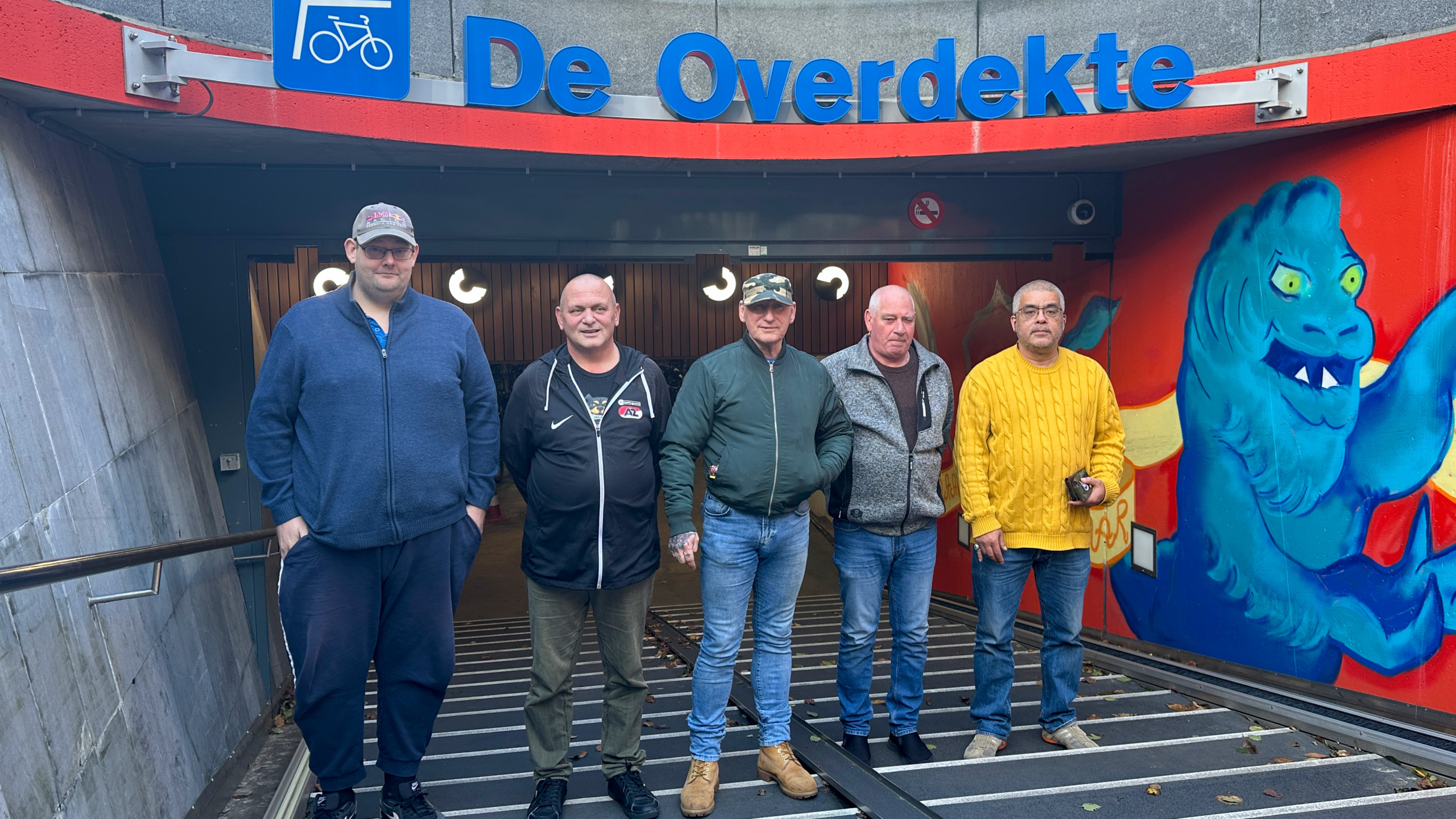 Vijf mannen poseren voor de ingang van 'De Overdekte', met een kleurrijk muurschildering en een bord met fietssymbool erboven, in een stedelijke omgeving.