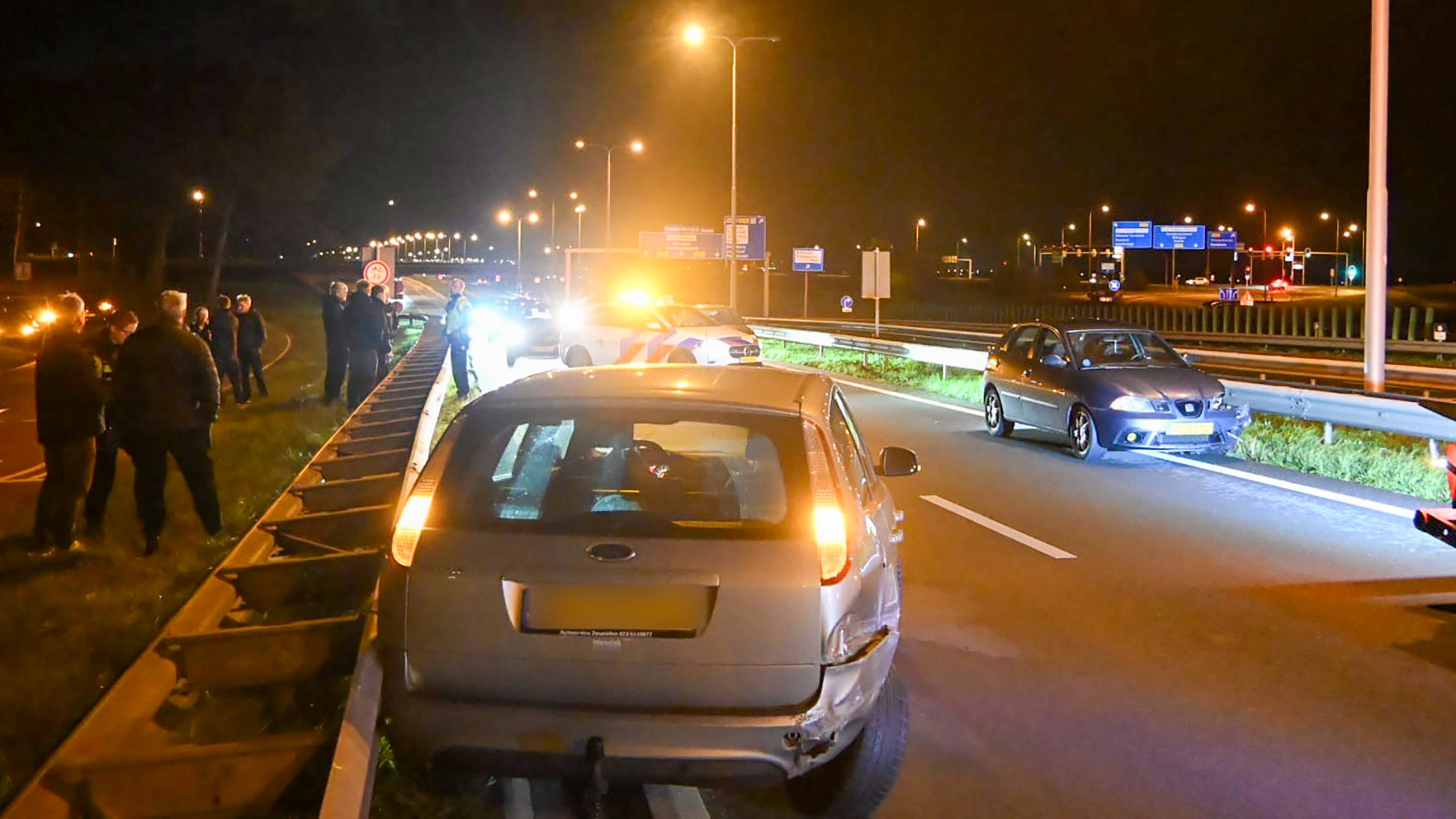 Verkeersongeval in de nacht op de snelweg, met meerdere auto's waarvan er één zichtbare schade heeft aan de achterkant. Politieauto's staan erbij met zwaailichten aan, en er zijn verschillende mensen langs de kant van de weg te zien.