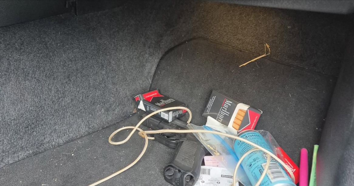Binnenkant van een kofferbak met pakjes sigaretten, een pistool, en andere spullen zoals een deodorantbus en papieren.