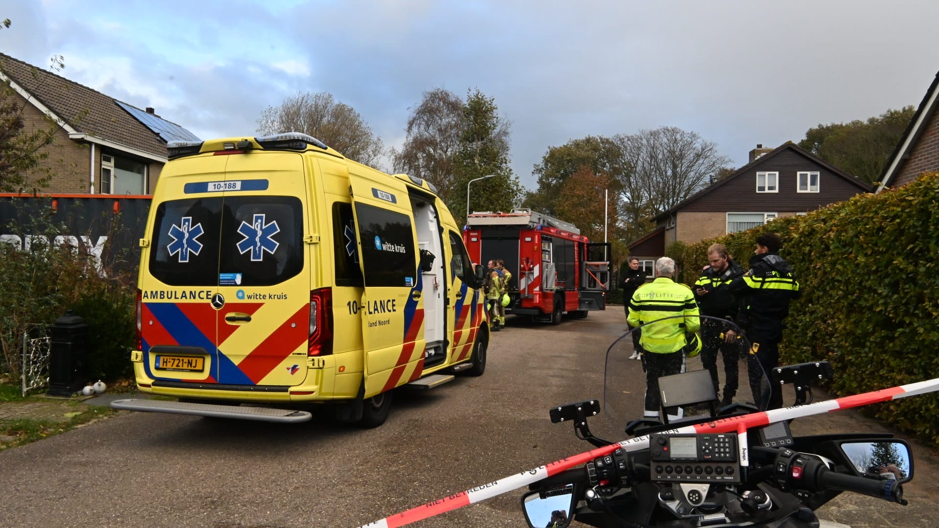 Een ambulance en brandweerwagen staan geparkeerd in een straat, omgeven door politieagenten die met elkaar praten, naast een afgezet gebied.