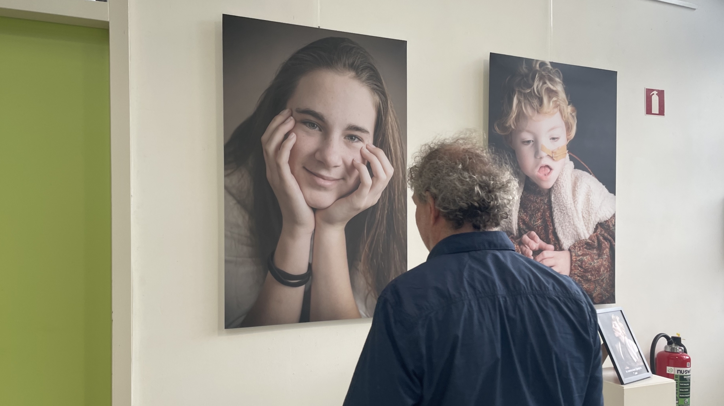 Een man bekijkt twee grote portretfoto's die aan een witte muur hangen, met links een foto van een lachend meisje en rechts een foto van een jongen met een pleister op zijn gezicht.