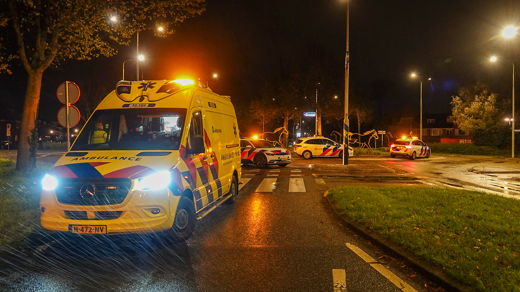 Een ambulance en meerdere politieauto's staan geparkeerd bij een rotonde op een natte straat tijdens de nacht, met oranje zwaailichten aan.