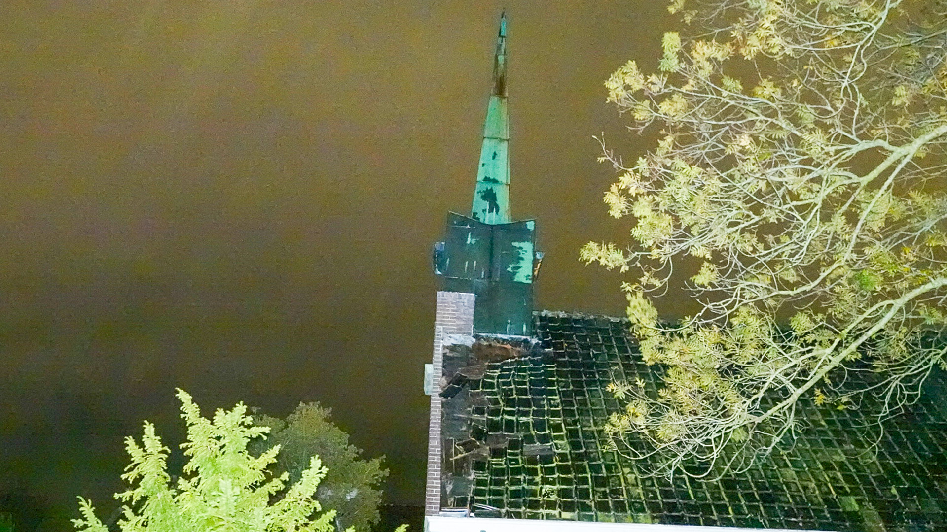 Een kerk met een beschadigde torenspits en dakpannen, omgeven door bomen, bij nacht.