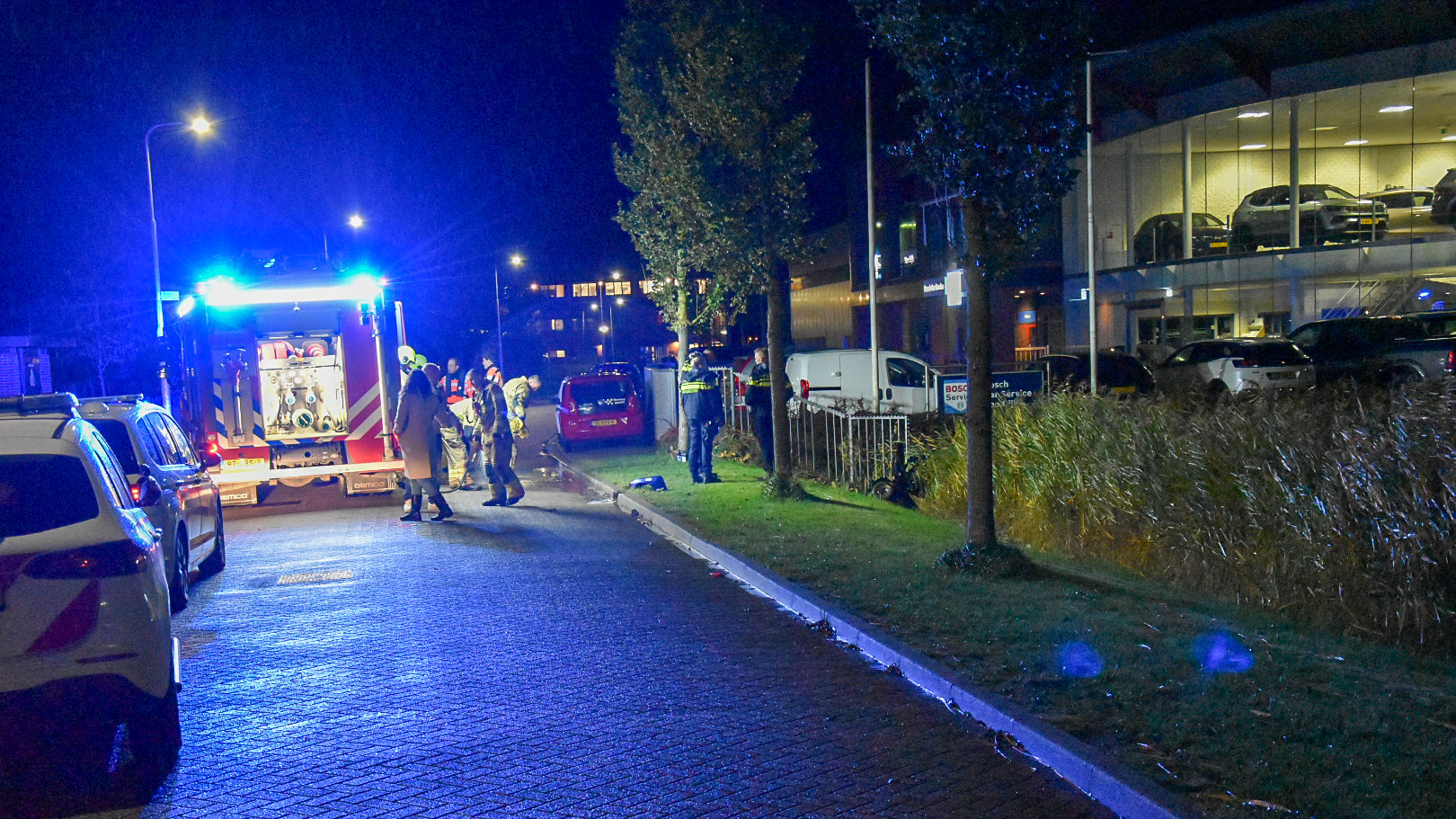 Brandweer en politie zijn aanwezig op een straat met blauwe lichten van een brandweerwagen die de omgeving verlichten, 's nachts geparkeerde auto's langs de weg en een showroom op de achtergrond.