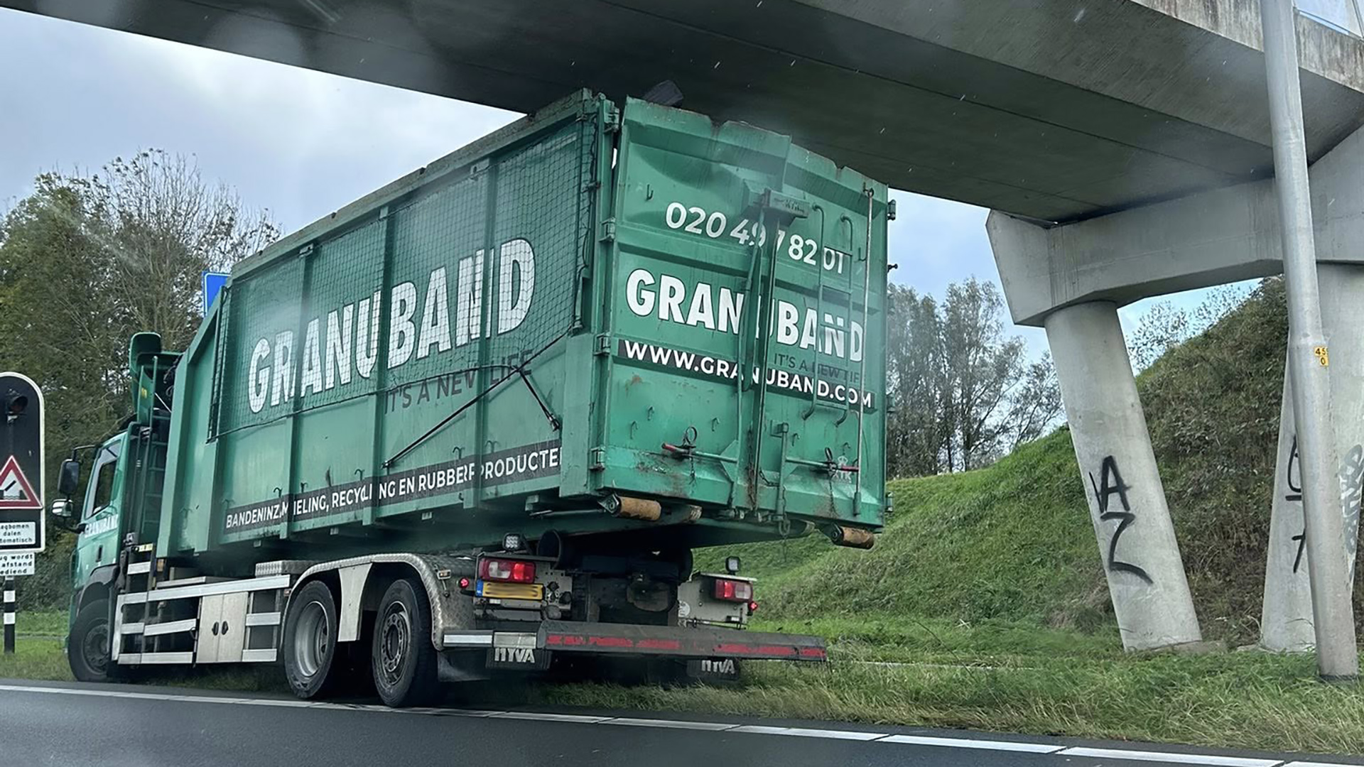 Groene vrachtwagen van het bedrijf Granuband stopt onder een viaduct naast een grashelling.