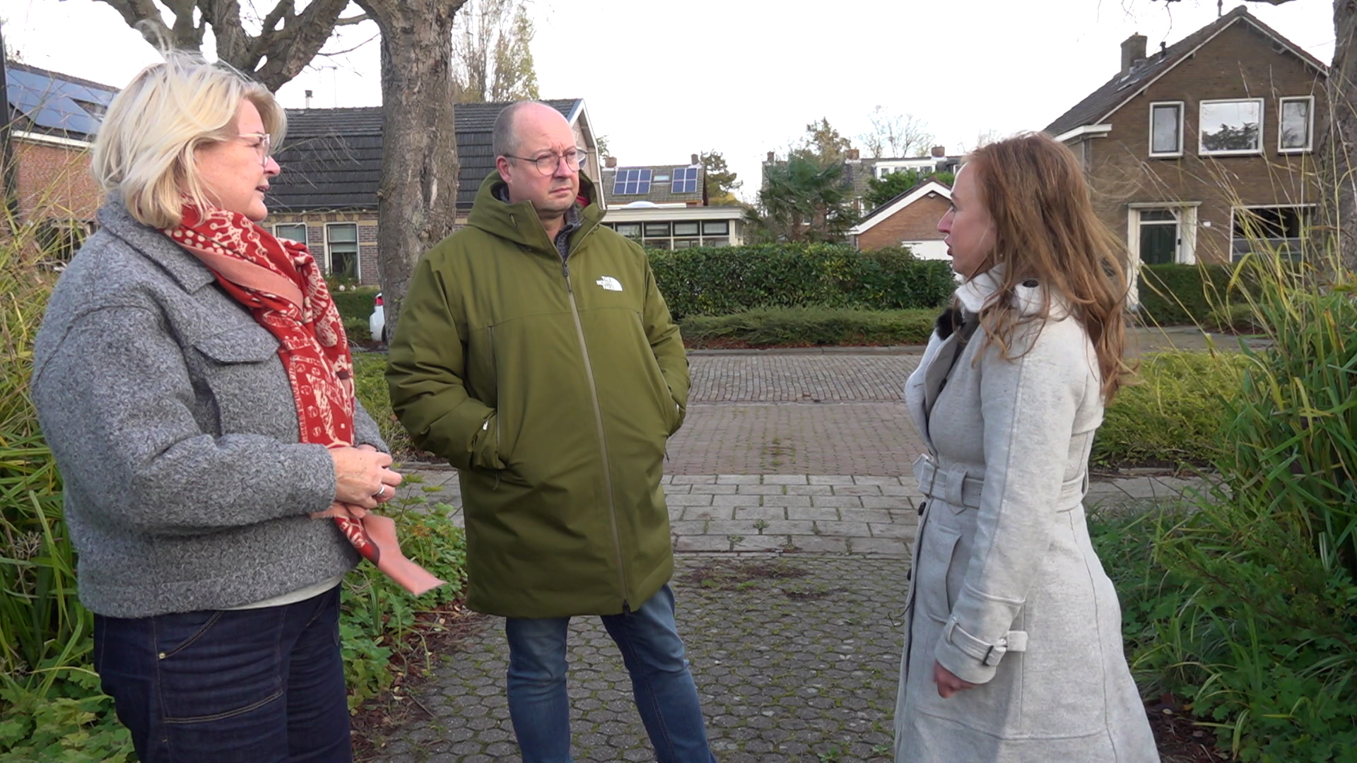 Drie mensen hebben een gesprek op een stoep in een woonwijk omgeven door huizen en groen.
