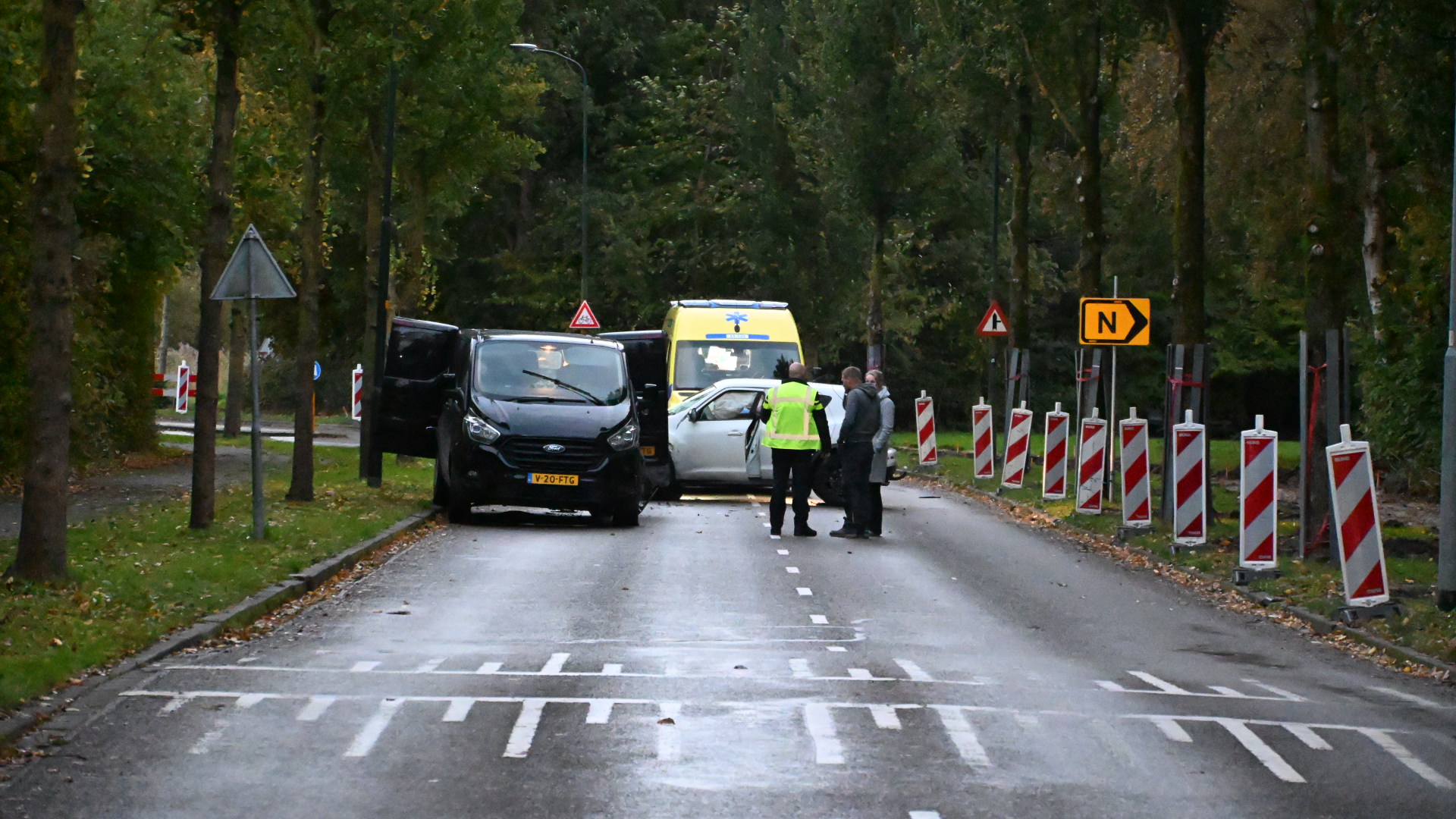 Twee voertuigen en een ambulance op een weg met wegversmalling en bordjes, omgeven door bomen. Meerdere mensen staan naast de voertuigen, een persoon in een geel hesje lijkt te communiceren.