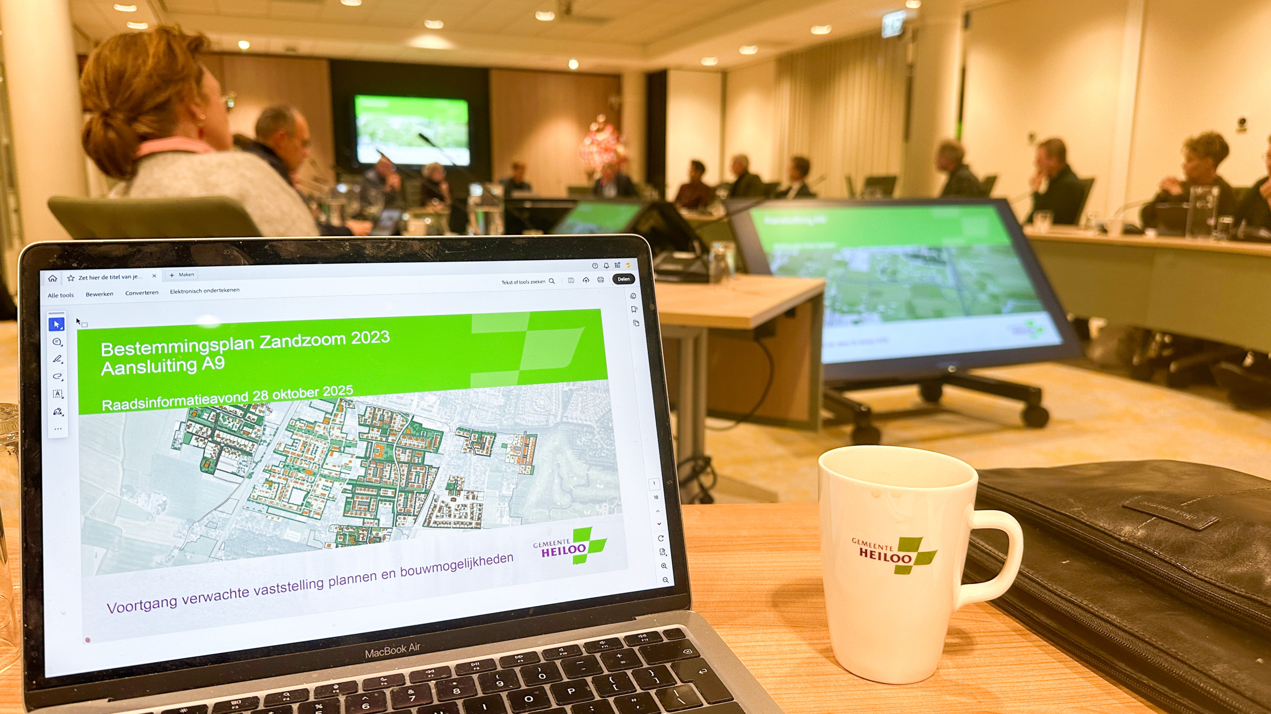 Vergaderzaal met een groep mensen zittend aan een tafel; op de voorgrond is een laptop te zien met een presentatie van het Bestemmingsplan Zandzoom 2023 en een koffiemok met het logo van gemeente Heiloo.