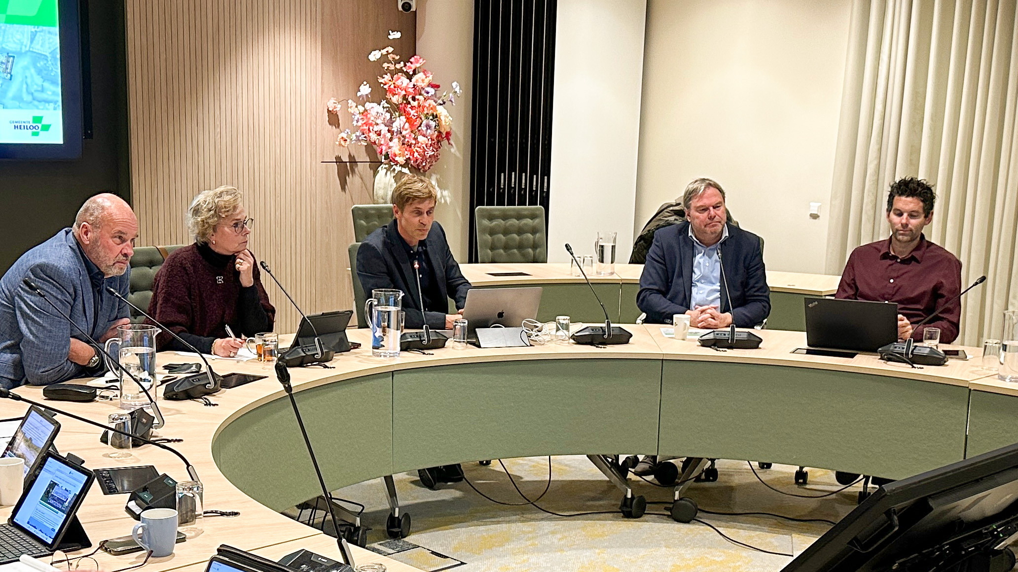 Vijf mensen zitten aan een ovale vergadertafel met laptops en notitieblokken, terwijl ze naar een scherm aan de muur kijken tijdens een vergadering.