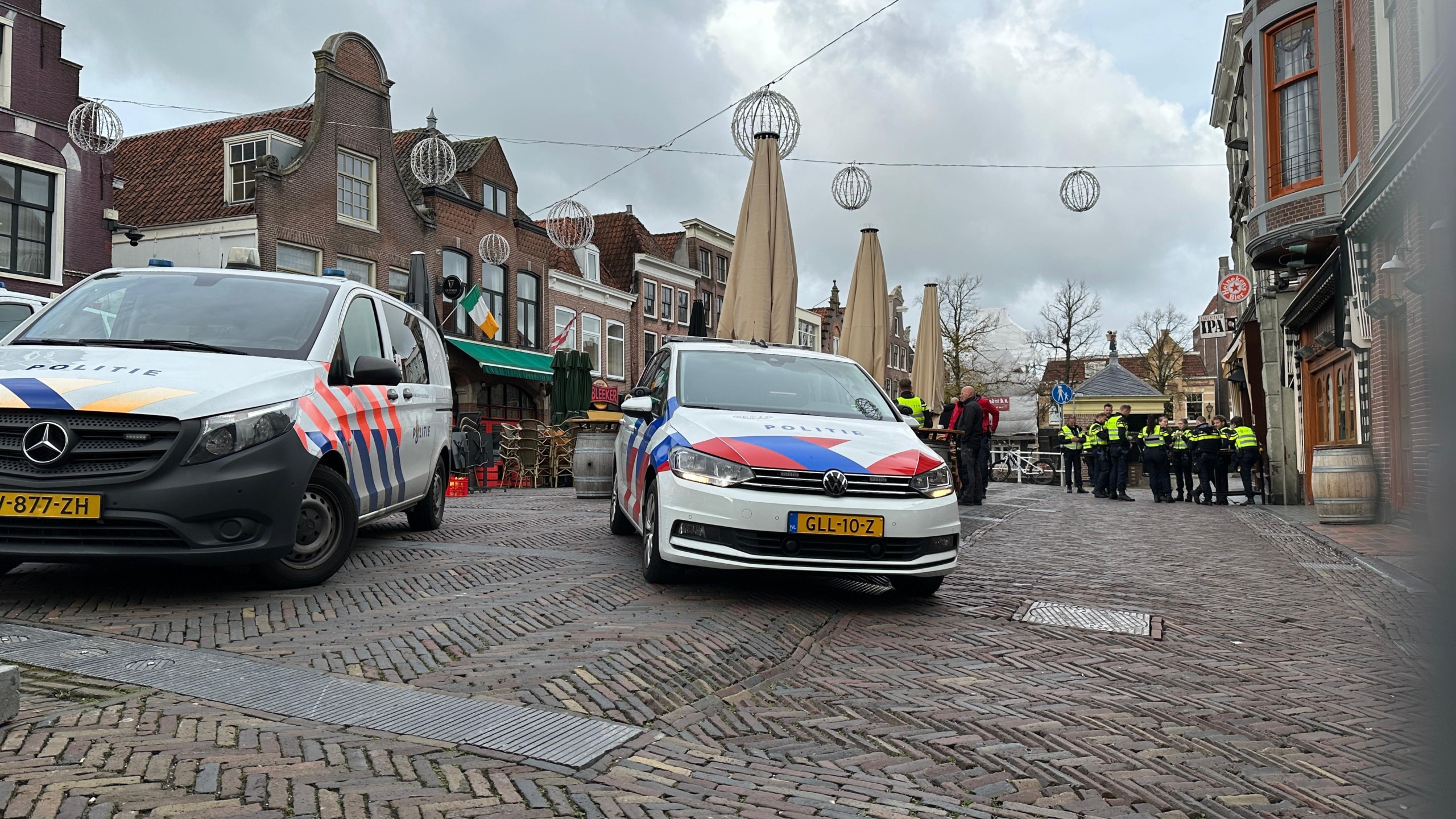 Straatbeeld met twee politieauto's op een geplaveide weg, omgeven door oude gebouwen en een groep politiemensen in uniform die verderop staan.