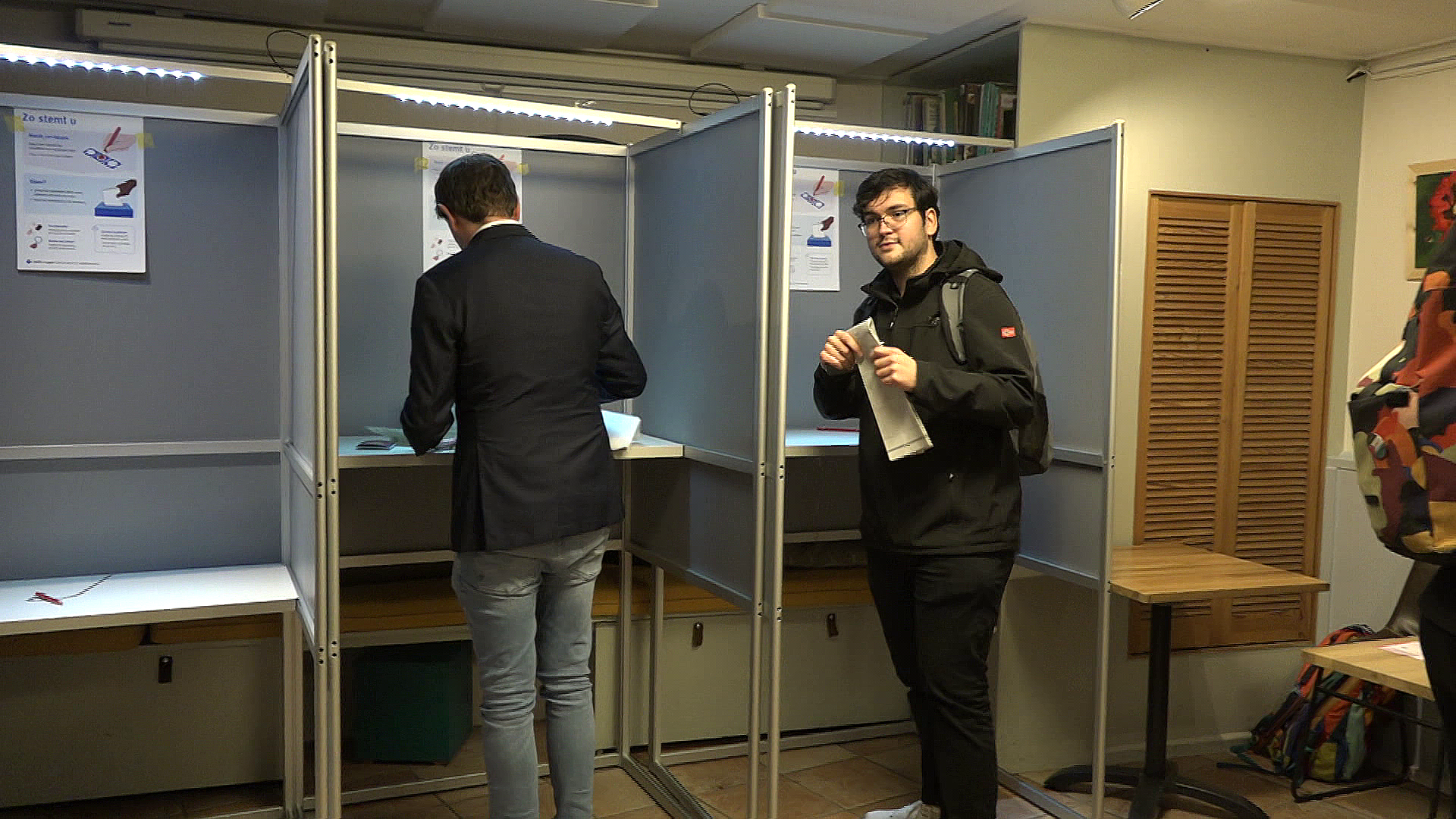 Twee mannen bij stemhokjes in een stemlokaal; de ene man staat in een hokje en de andere man houdt een stemformulier vast, kijkend naar de camera.