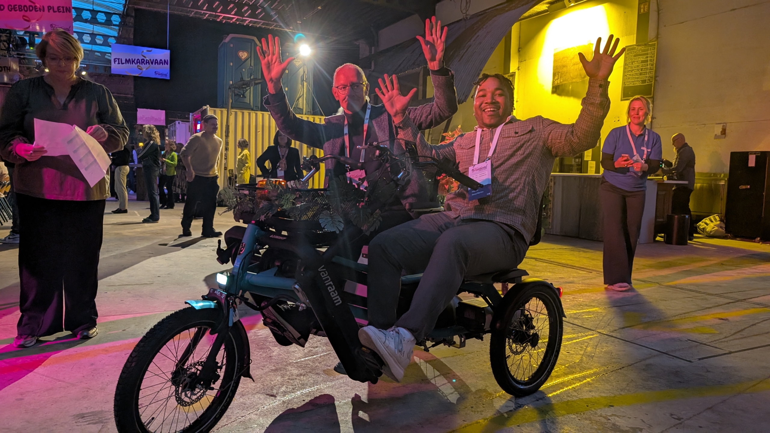 Twee mannen zitten op een driewieler met de handen opgeheven in een verlichte, industriële omgeving, omringd door andere mensen die staan en lopen. Borden en decoraties hangen in de achtergrond.