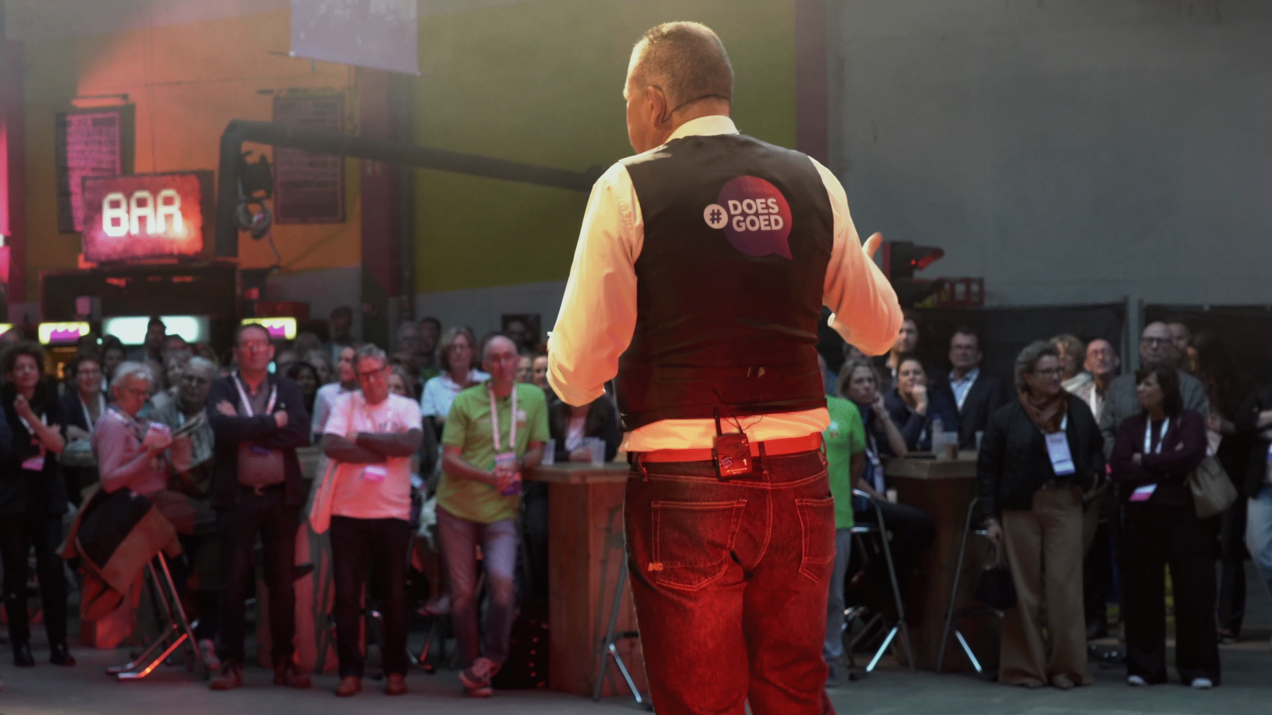 Een man spreekt op een podium voor een groep mensen bij een evenement, met een bar achtergrond.