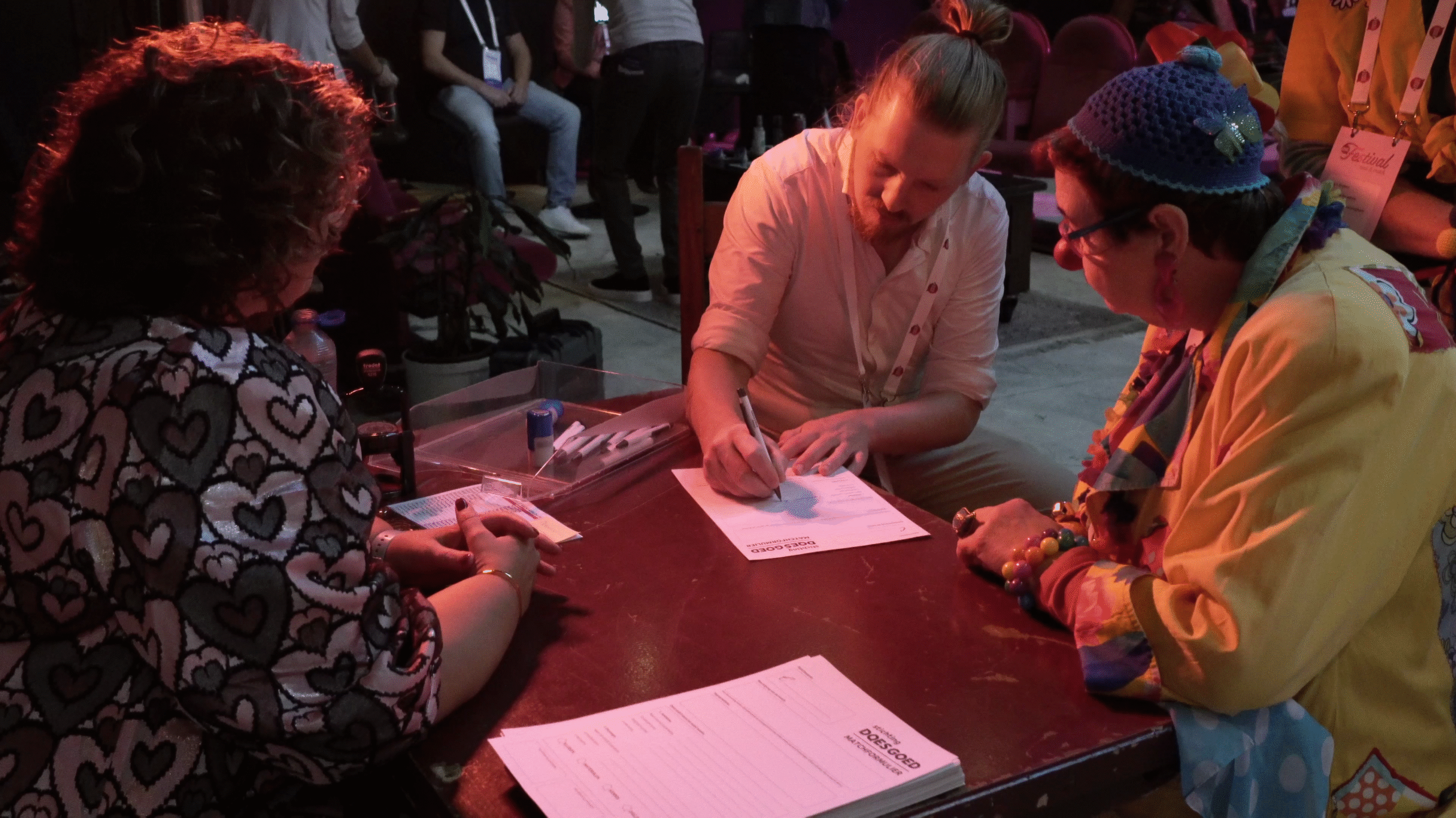 Een groep mensen zit aan een tafel waarvan één persoon een document ondertekent, terwijl anderen toekijken in een zaal met een informele sfeer.