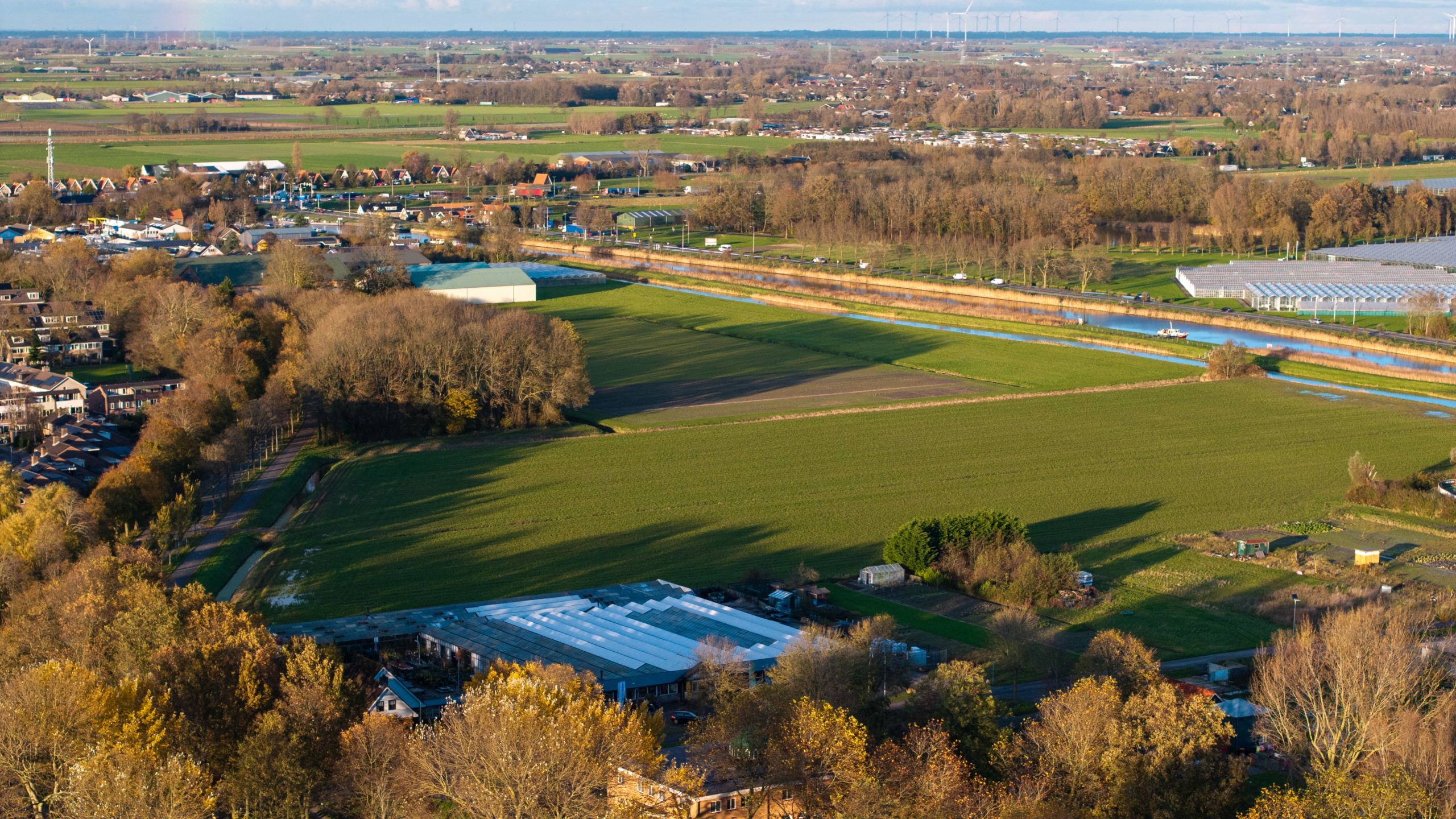 Luchtfoto van een Nederlands landschap met groene velden, een kanaal met een brug, en verspreide gebouwen, omringd door herfstbomen.