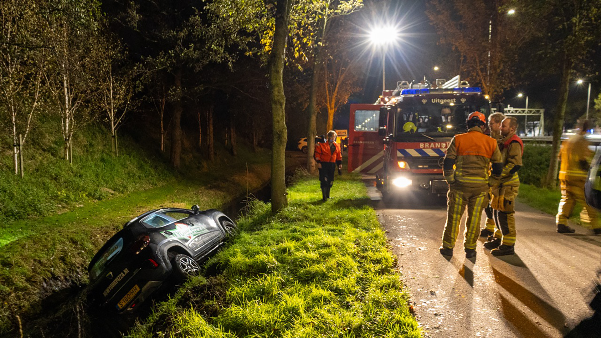Brandweerlieden staan naast een brandweerwagen op een verlichte weg, terwijl een auto schuin in een greppel ligt langs een grasstrook en bomen.