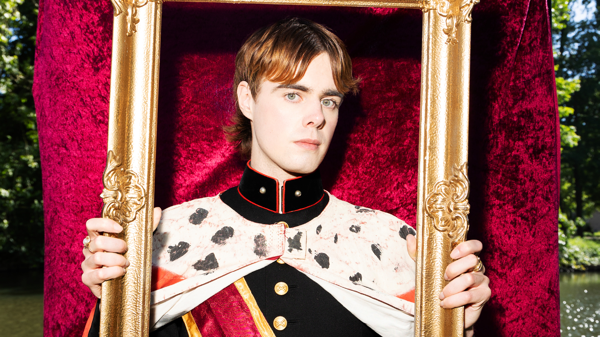 Een persoon in een koninklijk ogend kostuum met gouden knoppen en een cape met zwarte stippen, poseert terwijl hij een goudkleurige lijst vasthoudt tegen een rode achtergrond in een buitenomgeving.