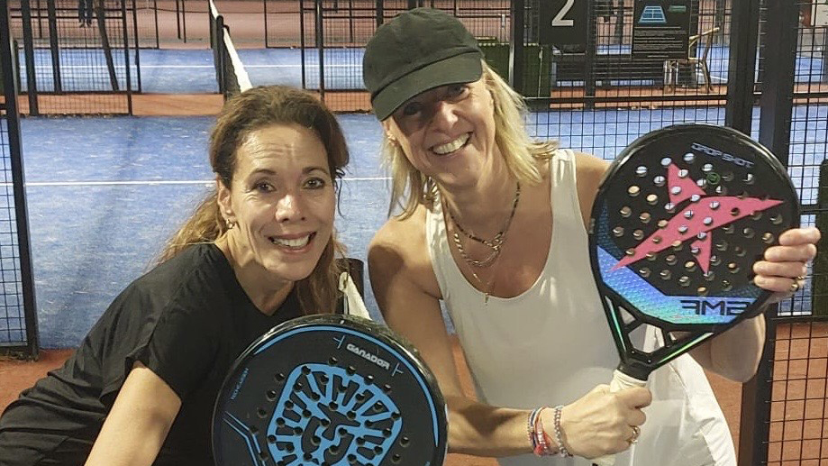 Twee lachende vrouwen op een padelbaan, elk met een padelracket zichtbaar in hun handen.