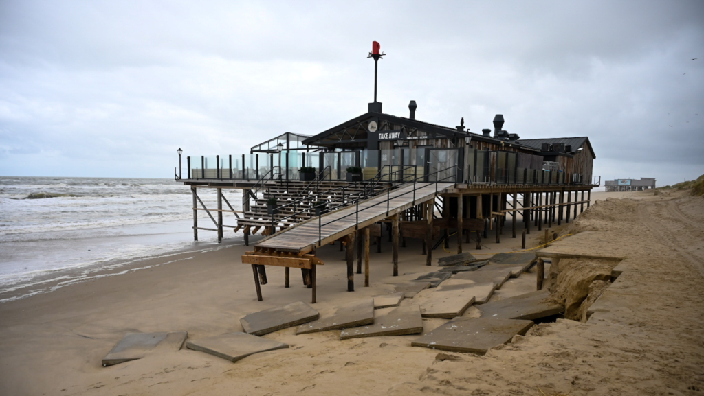 Strandpaviljoen op houten palen aan de kust, met een houten loopbrug en glazen balustrades, gelegen aan een zandstrand met zichtbare schade door erosie en een ruige zee op de achtergrond.