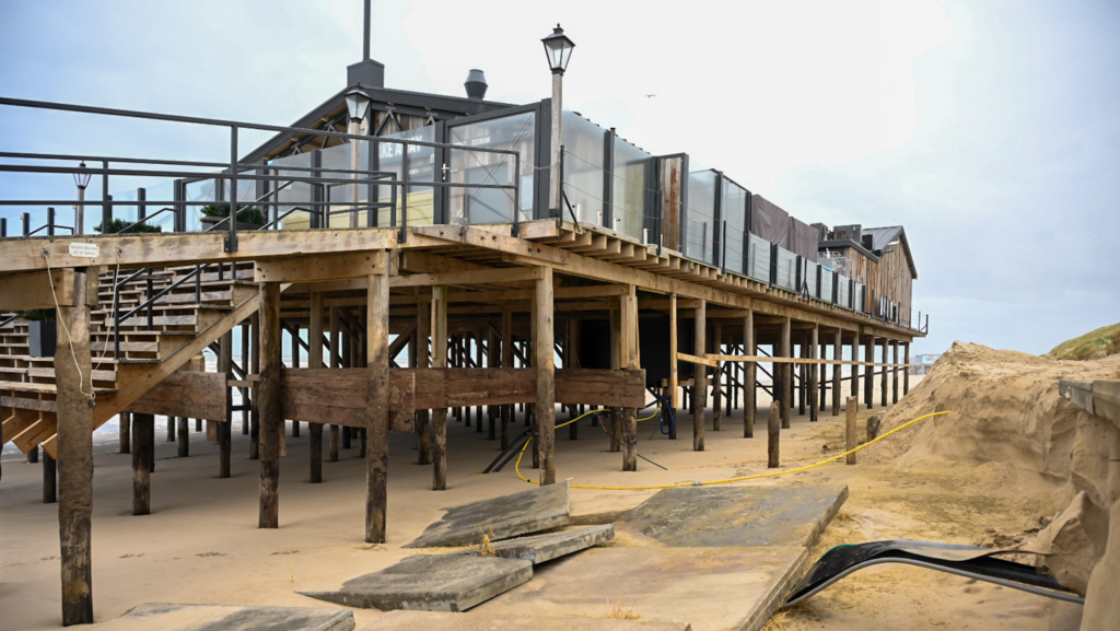 Een strandpaviljoen op houten palen aan de kust, met zand en losse betonnen platen op de voorgrond; de lucht is bewolkt.