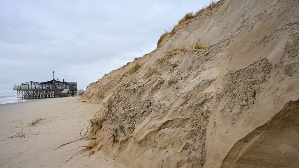 Een strand met hoge zandduinen aan de rechterkant en een gebouw op palen aan de linkerkant, gelegen aan de rand van de zee onder een bewolkte hemel.