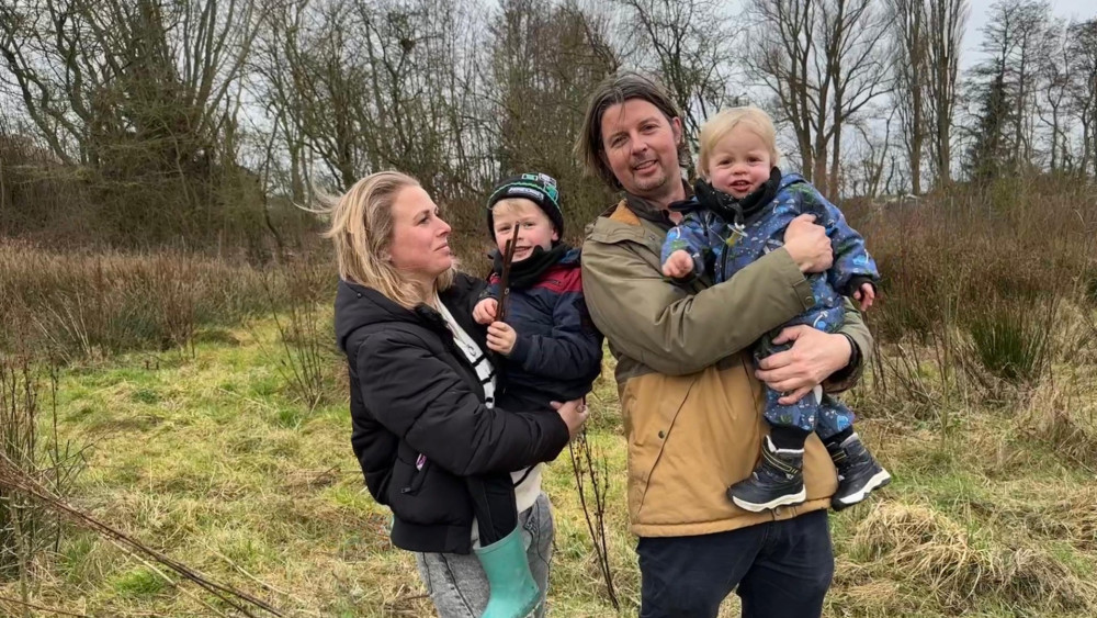 Een man en vrouw staan buiten op een grasveld, elk een kind vasthoudend; de vrouw draagt een donker jack en de man een beige jas.