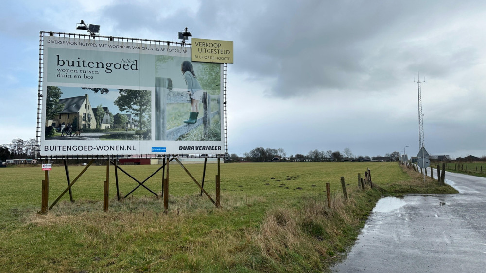 Een groot reclamebord met daarop de tekst 'Buitengoed, wonen tussen duin en bos' en afbeeldingen van een huis en een kind op een hek. Een lege grasveld en landweg zijn op de achtergrond te zien, onder een bewolkte lucht.