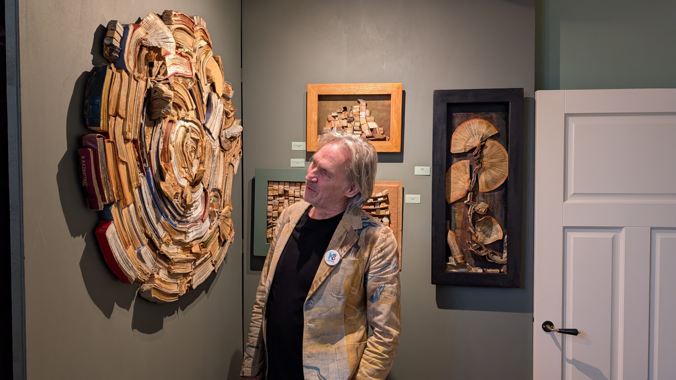 Een man kijkt naar een kunstwerk aan de muur gemaakt van boeken en papier, met andere kunstwerken op de achtergrond in een galerij.
