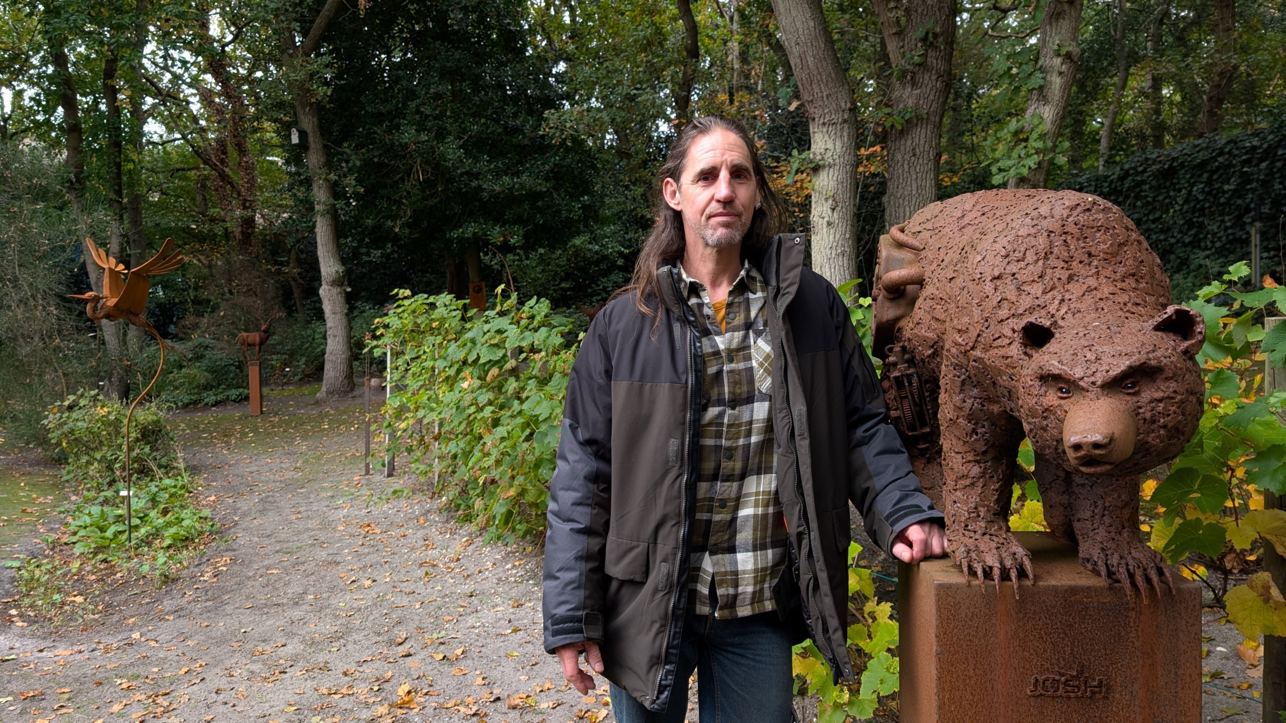 Een man staat buiten naast een metalen berenbeeld in een bosrijke omgeving.