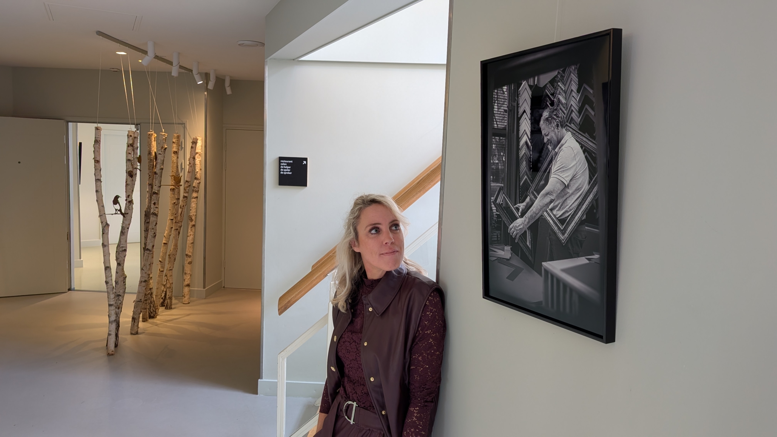 Vrouw leunt tegen een muur naast een ingelijste zwart-wit foto van een man in een galerie, met op de achtergrond opgehangen berkenboomstammen.