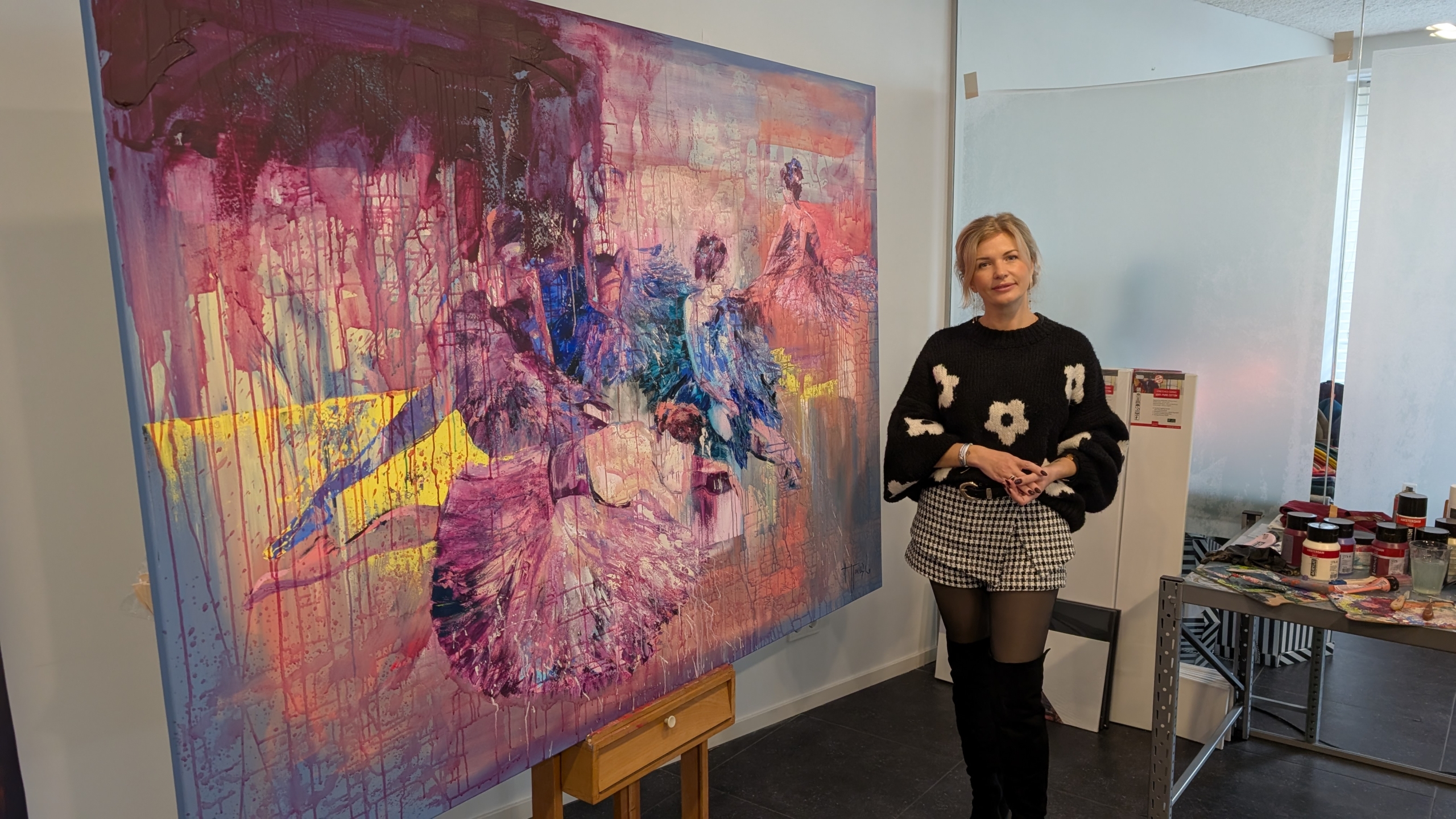 Een vrouw staat naast een groot kleurrijk schilderij met abstracte figuren en felgekleurde penseelstreken. Ze draagt een zwart trui met witte bloemmotieven en geruite rok, en staat in een kunststudio met potten verf op een tafel op de achtergrond.