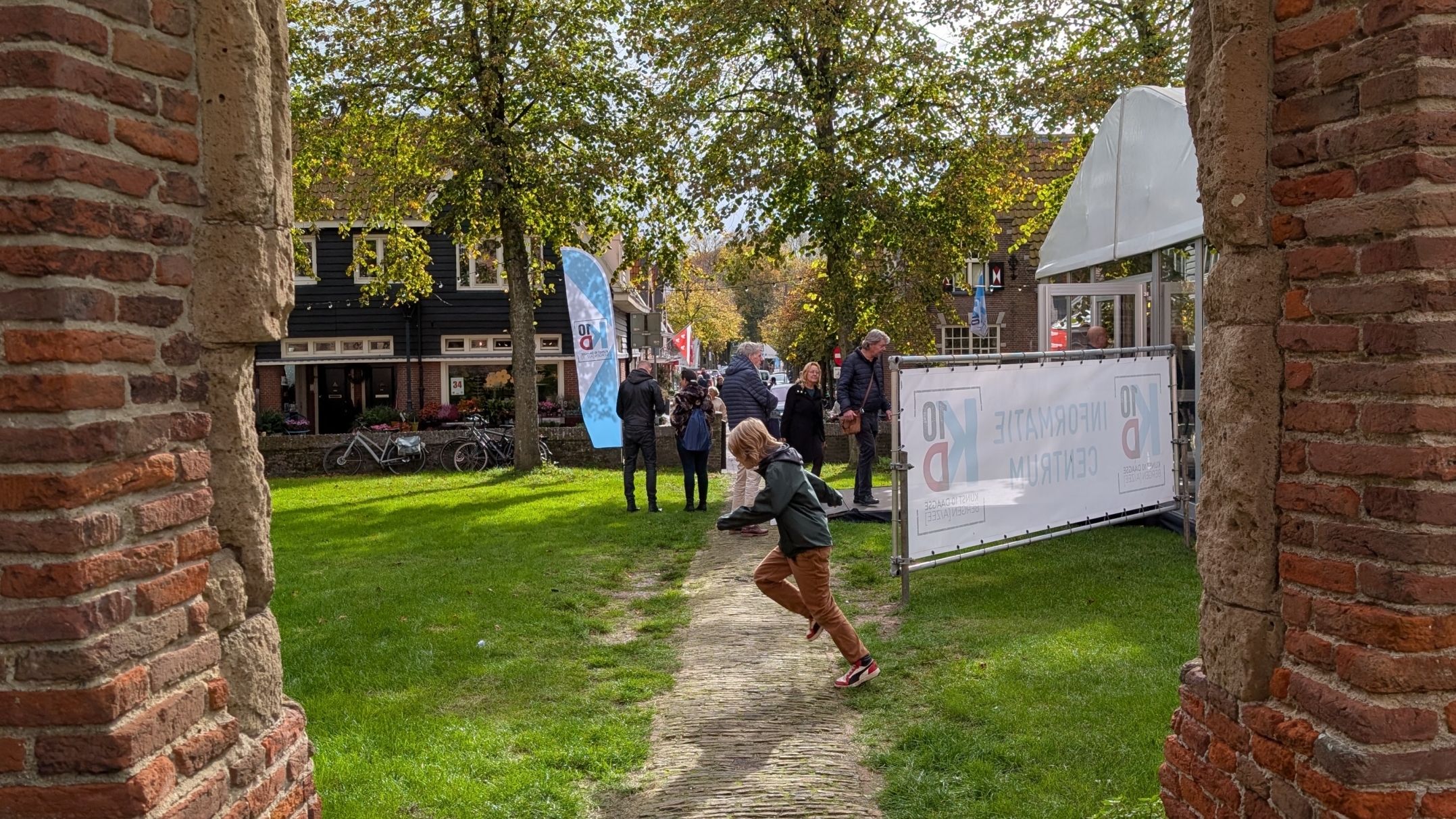 Een kind rent over een stenen pad tussen twee rode bakstenen muren met groen gras en een informatiecentrum op de achtergrond, omgeven door bomen en mensen.