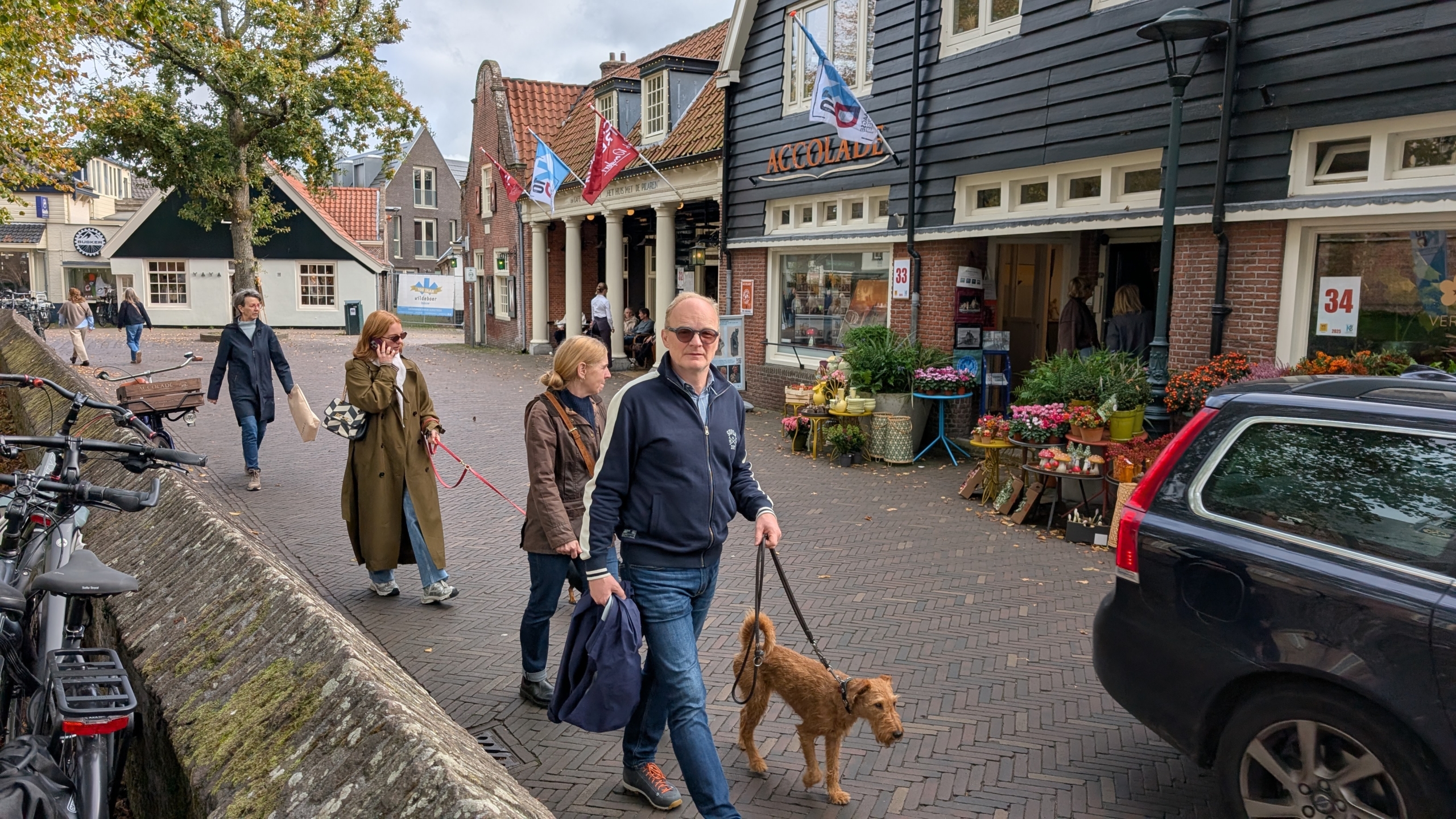 Een man met een hond aan de lijn loopt voor een bloemenwinkel aan een straat met geplaveide stenen, terwijl verschillende mensen voorbijlopen en fietsen aan de linkerkant zijn geparkeerd.