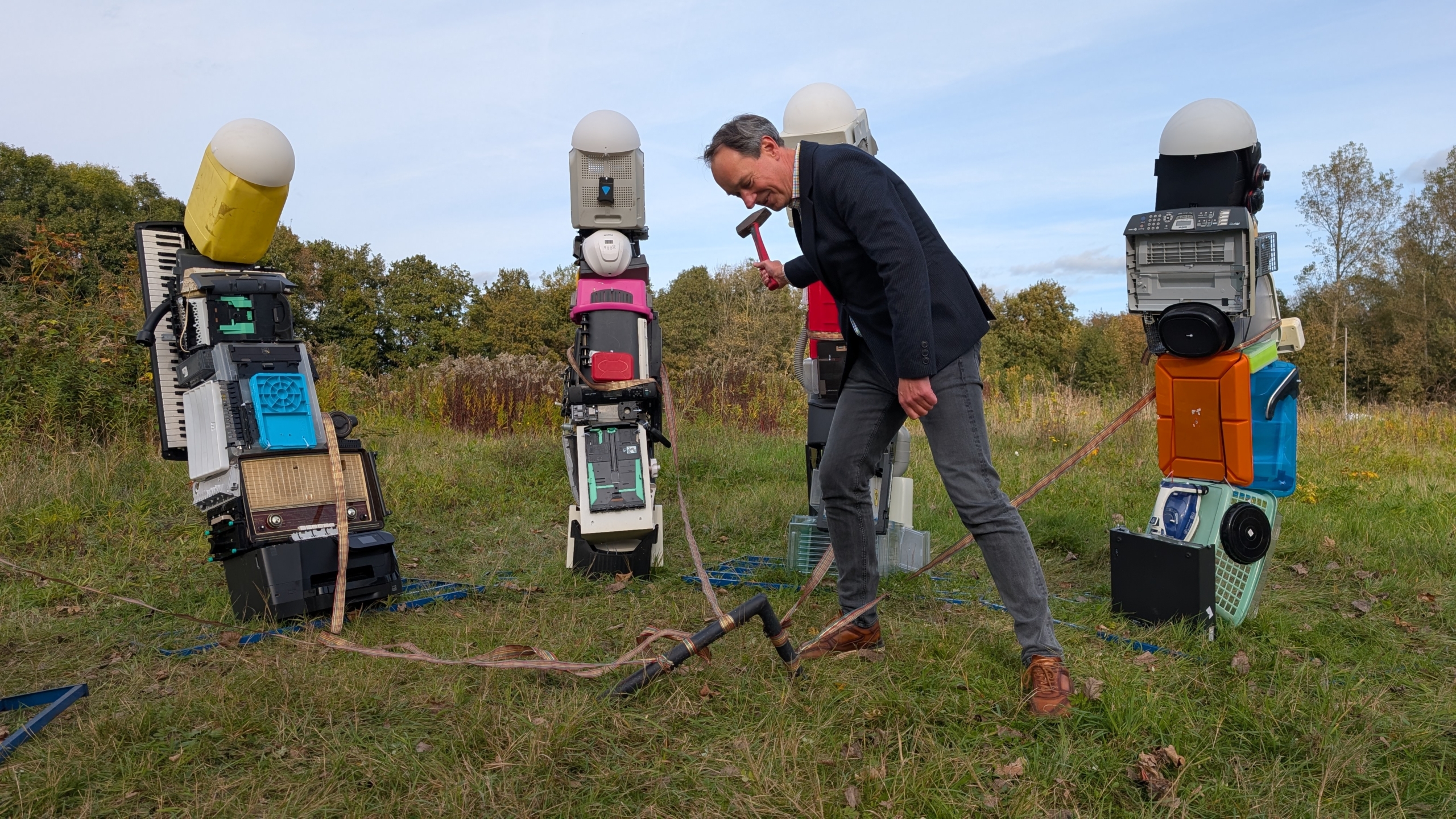 Een man in een jasje gebruikt een hamer om een paal in de grond te slaan naast kunstwerken gemaakt van gerecyclede elektronische apparaten op een grasveld.