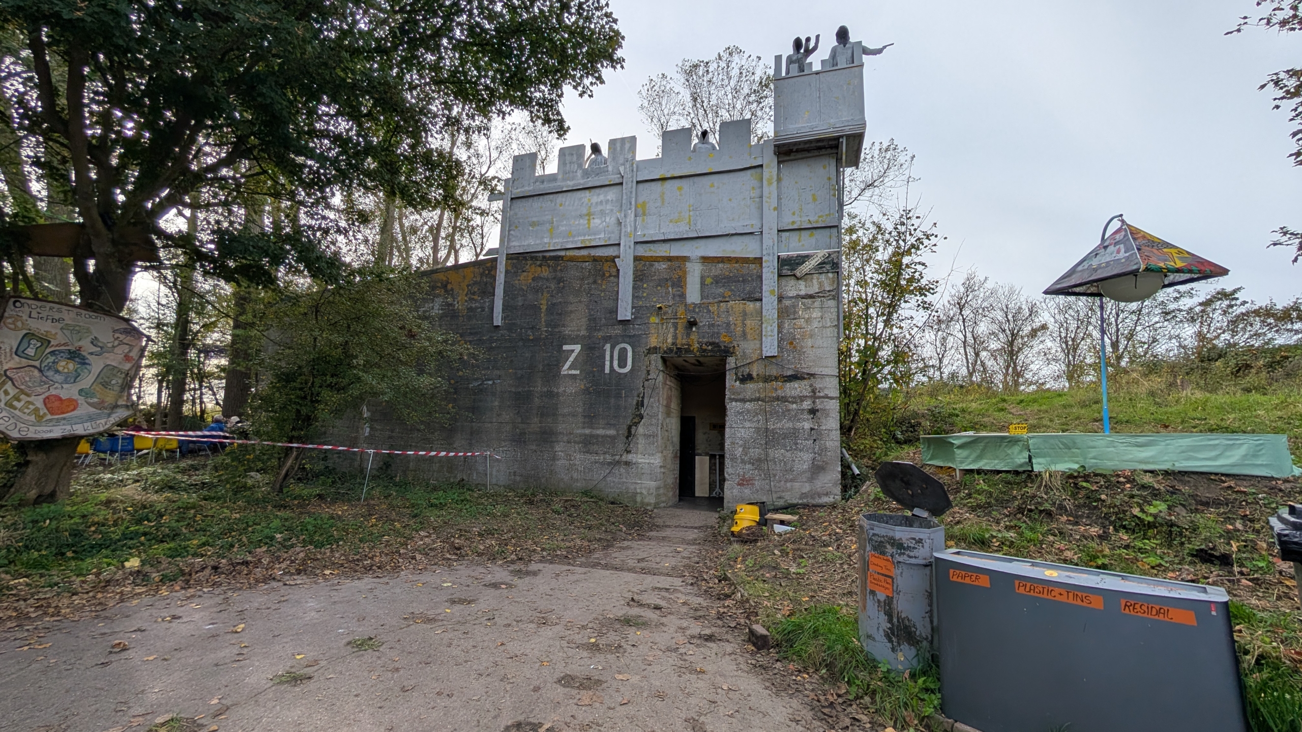 Een betonnen bunker met het nummer Z 10 onder een bewolkte lucht, met een torenuitbouw en beeldhouwwerken erbovenop. Links staan bomen en hangt een bord met tekeningen, rechts een kleurrijk, paraplu-vormig object nabij afvalbakken met labels voor verschillende materialen.