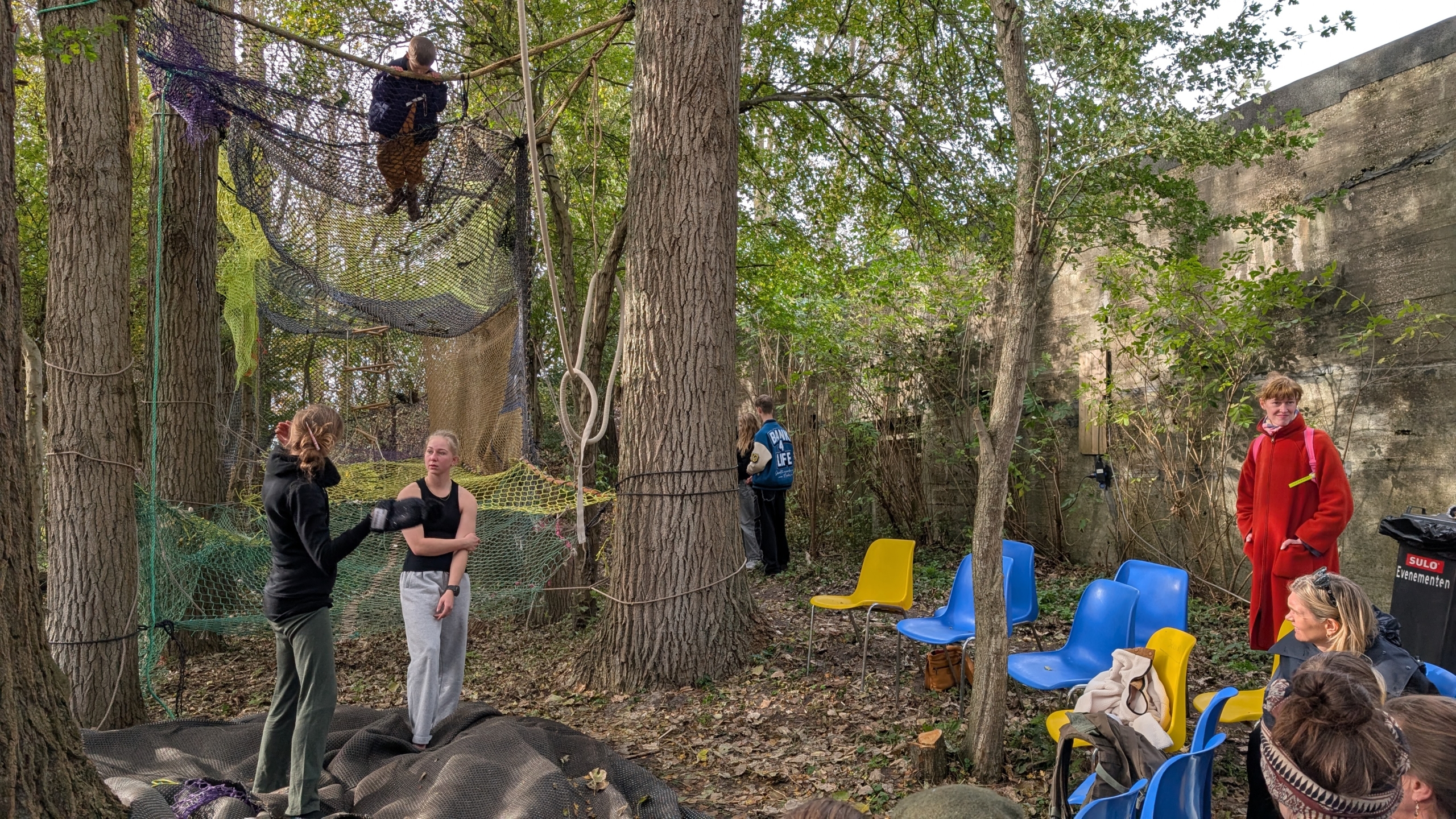 Een groep mensen in een bosrijke omgeving met netten tussen de bomen waar iemand op klimt. Er zijn kleurrijke stoelen, een persoon in een rode jas en anderen die toekijken of praten. De omgeving lijkt een speelse en creatieve ruimte.