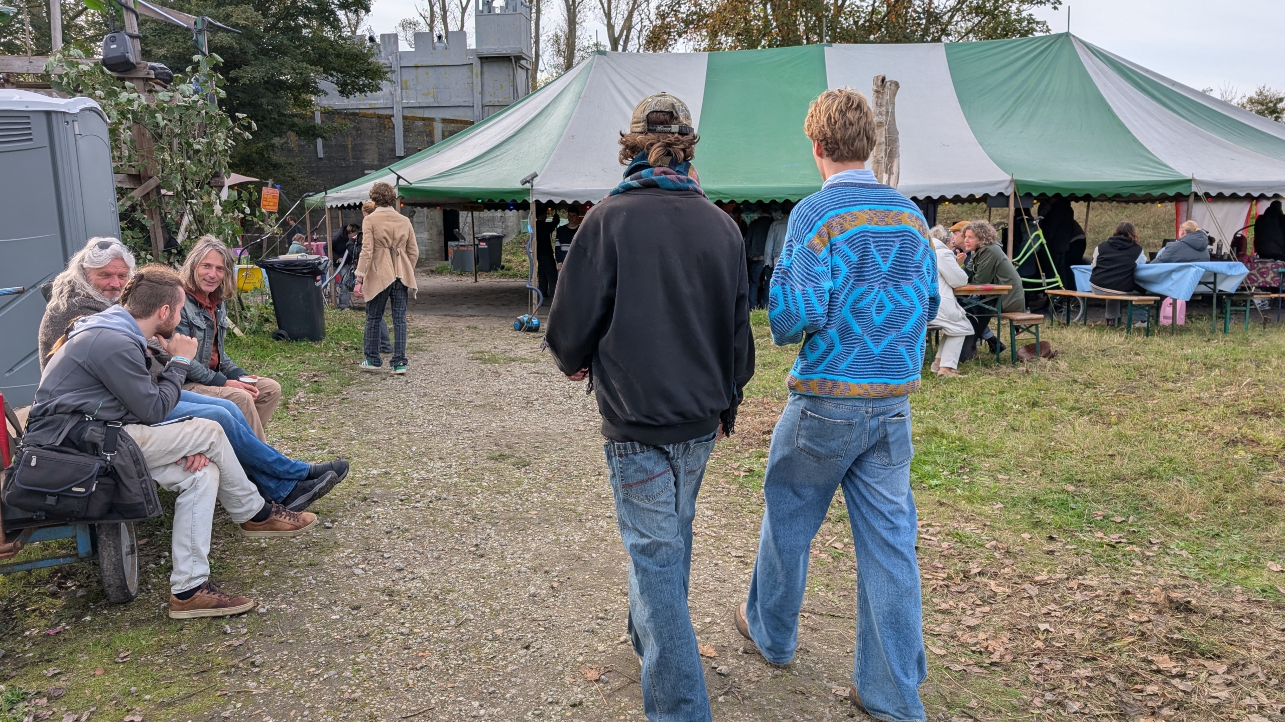 Een buitenlocatie met een feesttent, waar mensen aan tafels zitten en ontspannen onder een groen-wit gestreepte tent. Op de voorgrond lopen twee personen bij de tent weg, terwijl links drie mensen op een bank zitten praten. Het terrein is bedekt met gras en bladeren.